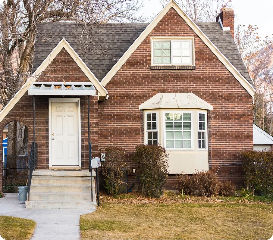 Brick house with white door, steps, bay window, and leafless bushes in front yard.