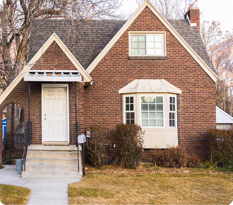 Brick house with white door, steps, bay window, and leafless bushes in front yard.