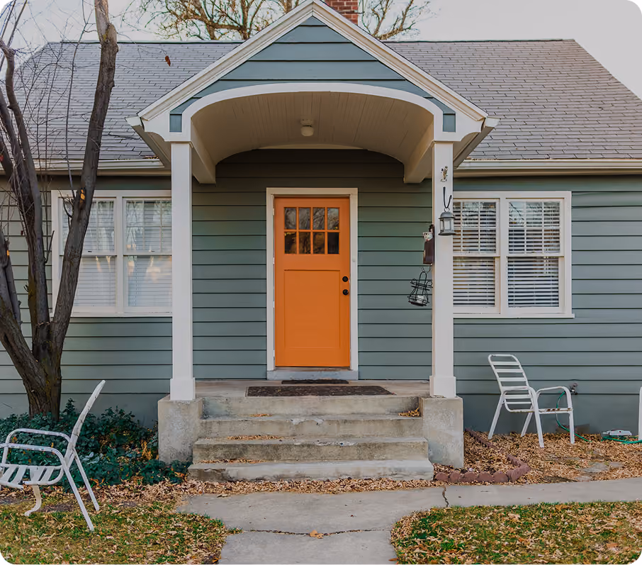 Front porch of a gray house with an orange door, two white metal chairs, and fall leaves on the ground.
