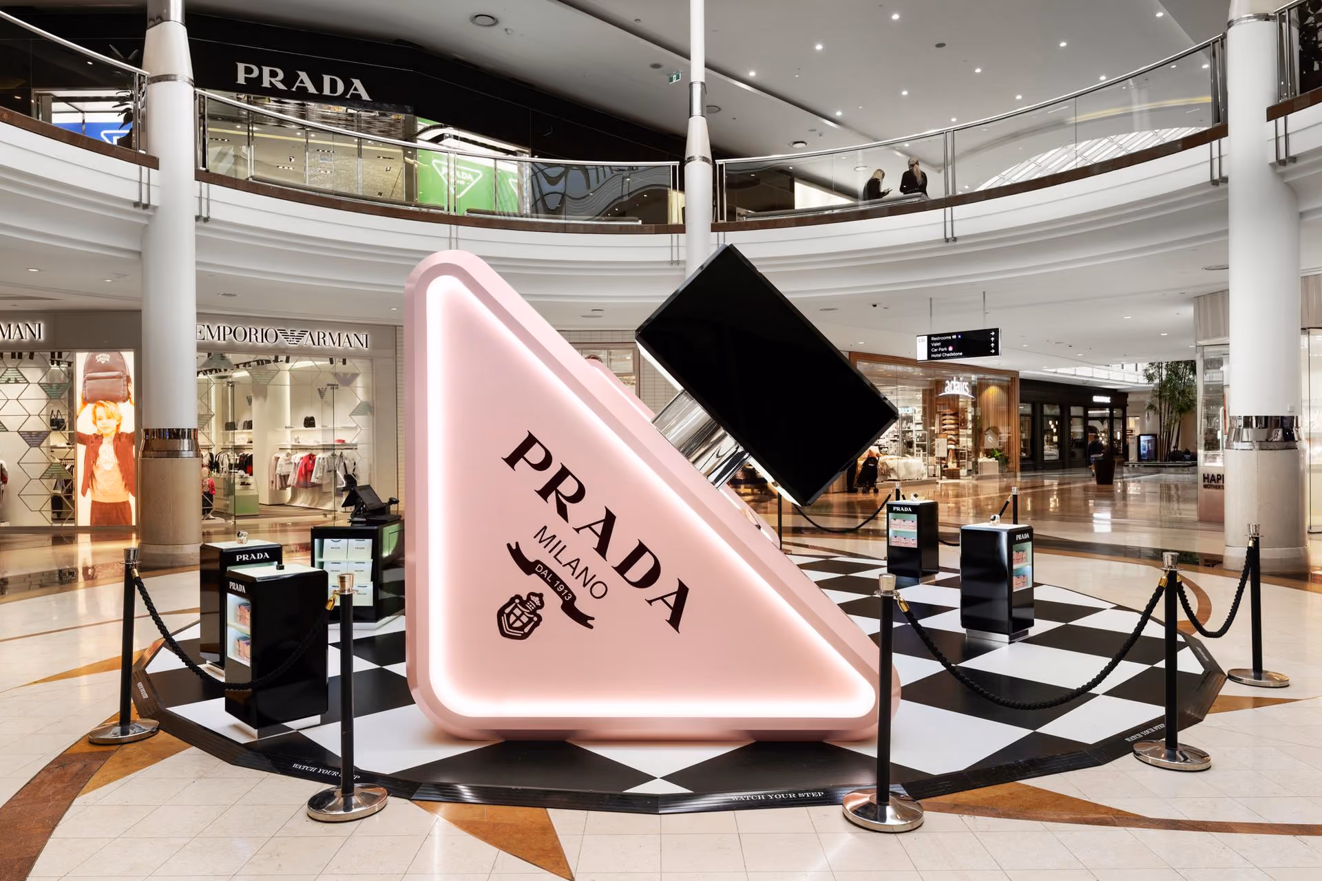 Giant Prada perfume bottle display in a shopping mall with Emporio Armani and Prada stores nearby.