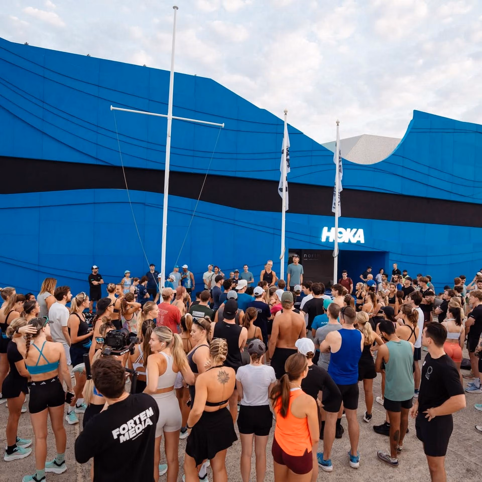 Crowd of people in athletic wear gathered outside a building with a blue wave design and HOKA sign.