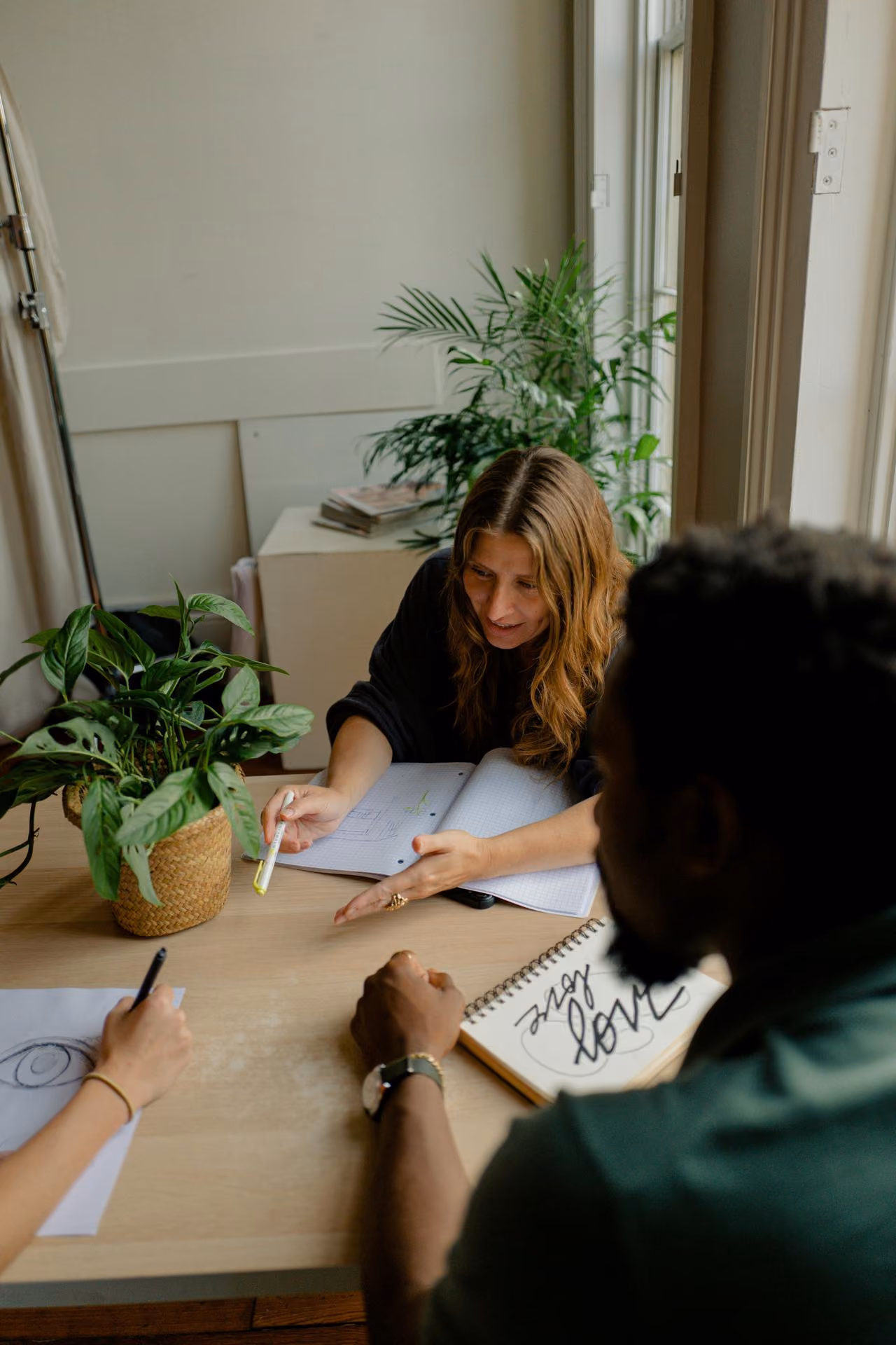 Woman with wavy hair points at notebook while two others sketch and write at table with a potted plant.