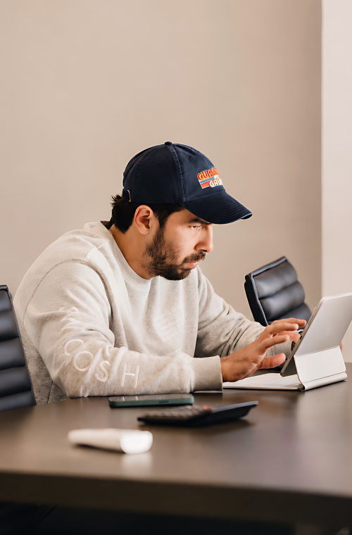 Man wearing a baseball cap and sweatshirt using a tablet at a table with chairs.