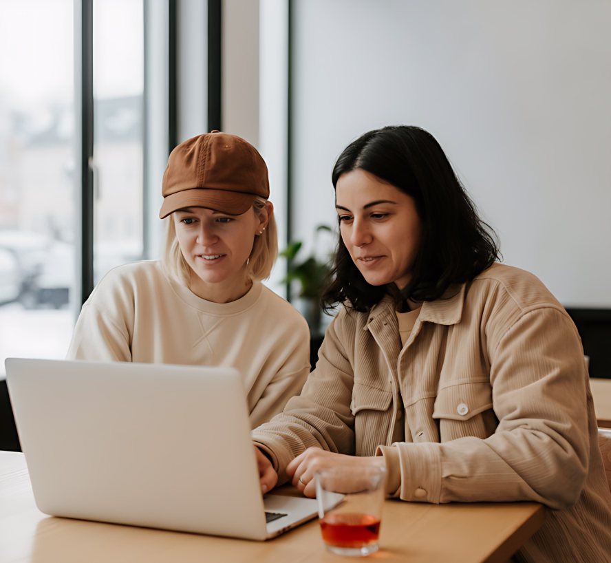 Two women sitting at a table looking at a laptop screen together in a bright room.