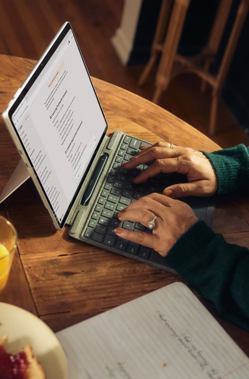 Person typing on a tablet with a keyboard on a wooden table, with a notebook and food nearby.