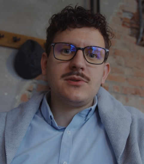 Man with glasses, curly hair, and mustache wearing a blue shirt and gray sweater indoors.