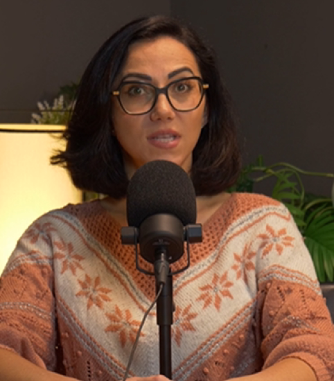 Woman with glasses speaking into a microphone wearing a patterned sweater in an indoor setting.
