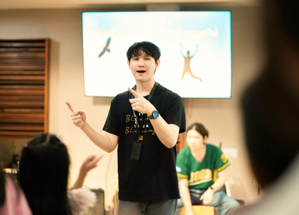 Young man speaking and gesturing in front of an audience with another person seated behind him.