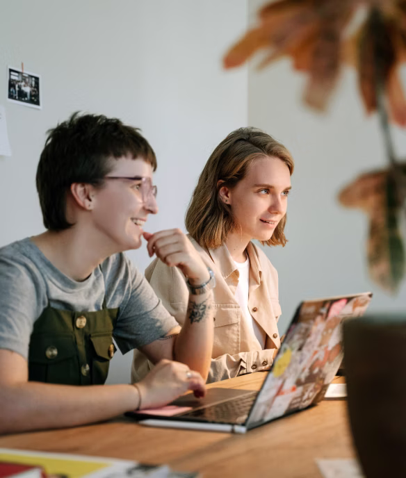 Two young people smiling and working together on a laptop at a wooden table indoors.