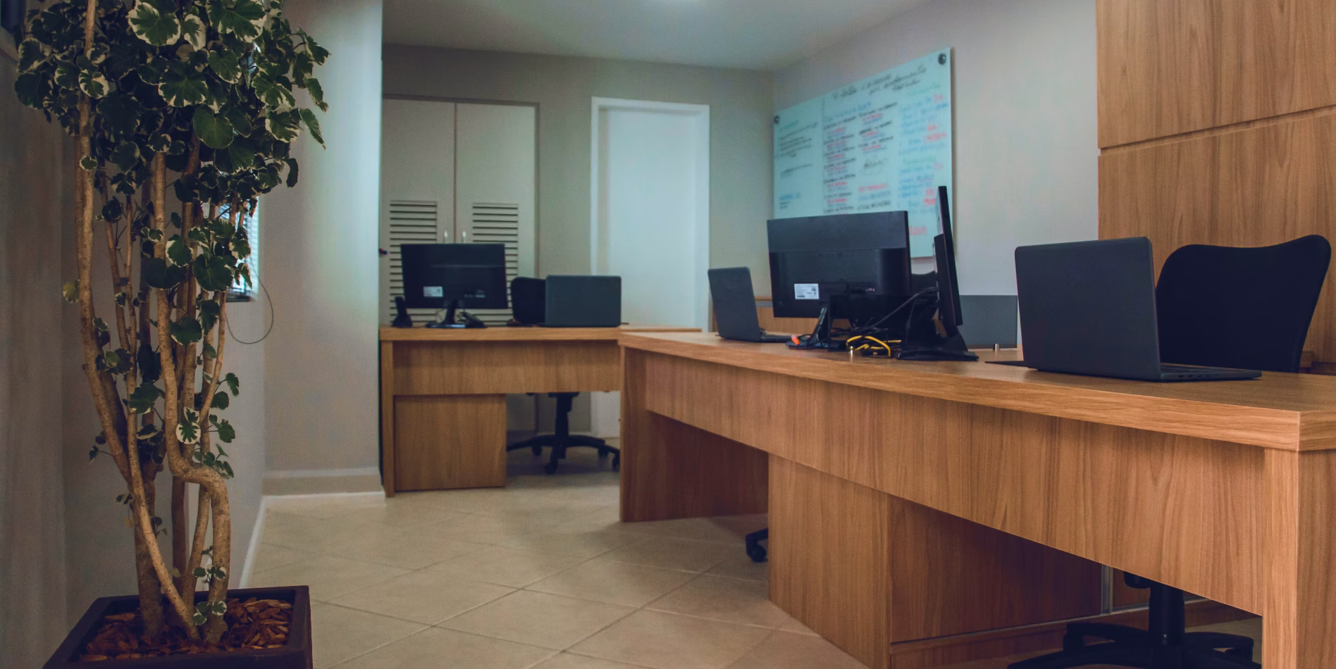 Modern office with wooden desks, laptops, monitors, chairs, indoor plant, and a whiteboard in the background.