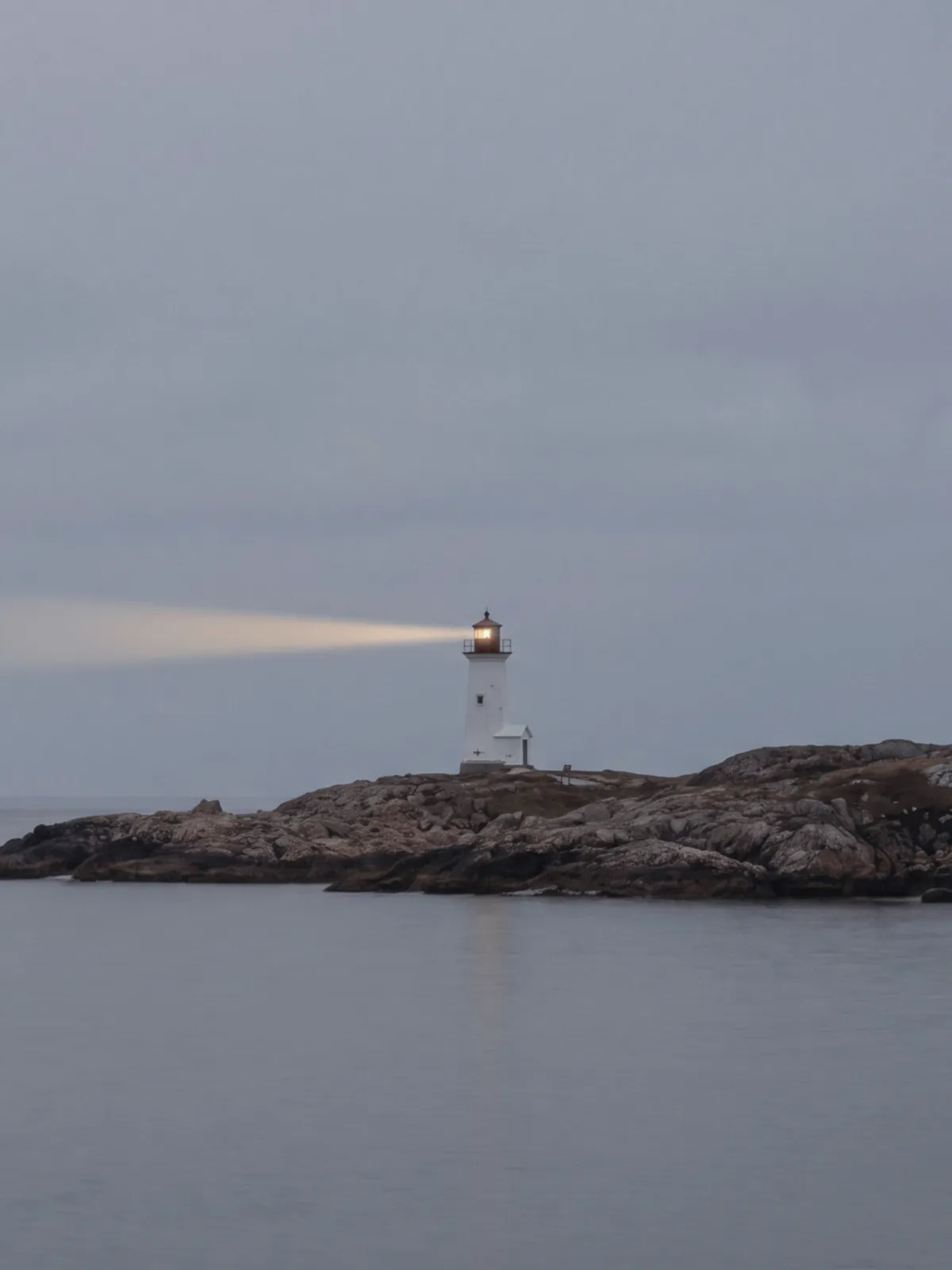 A lighthouse beam reaching across a calm grey sea at dusk