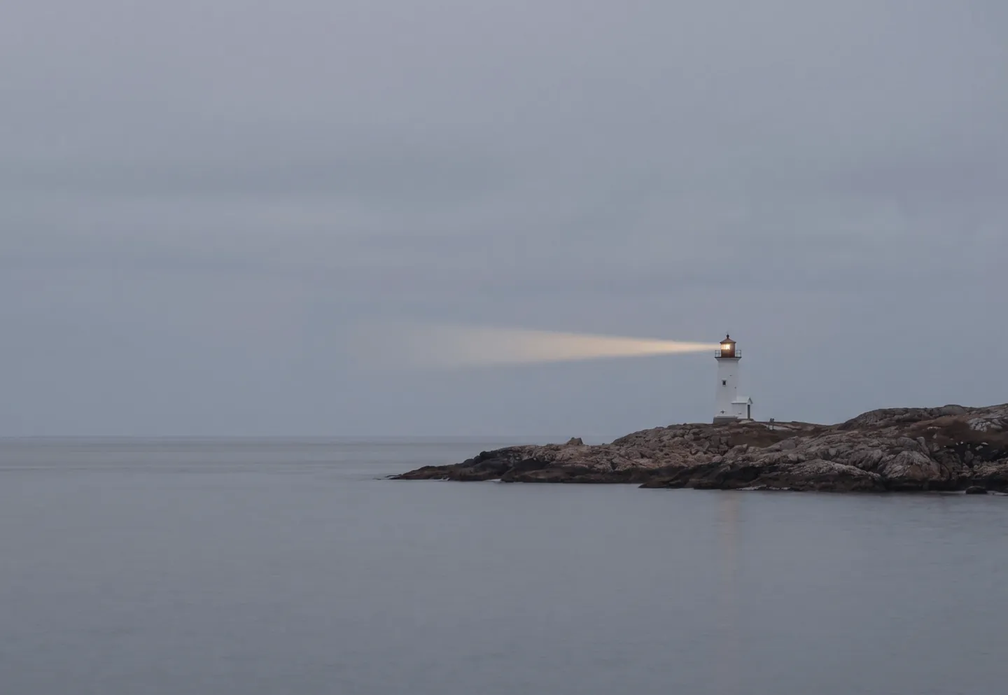 A lone lighthouse shining across a calm grey sea at dusk