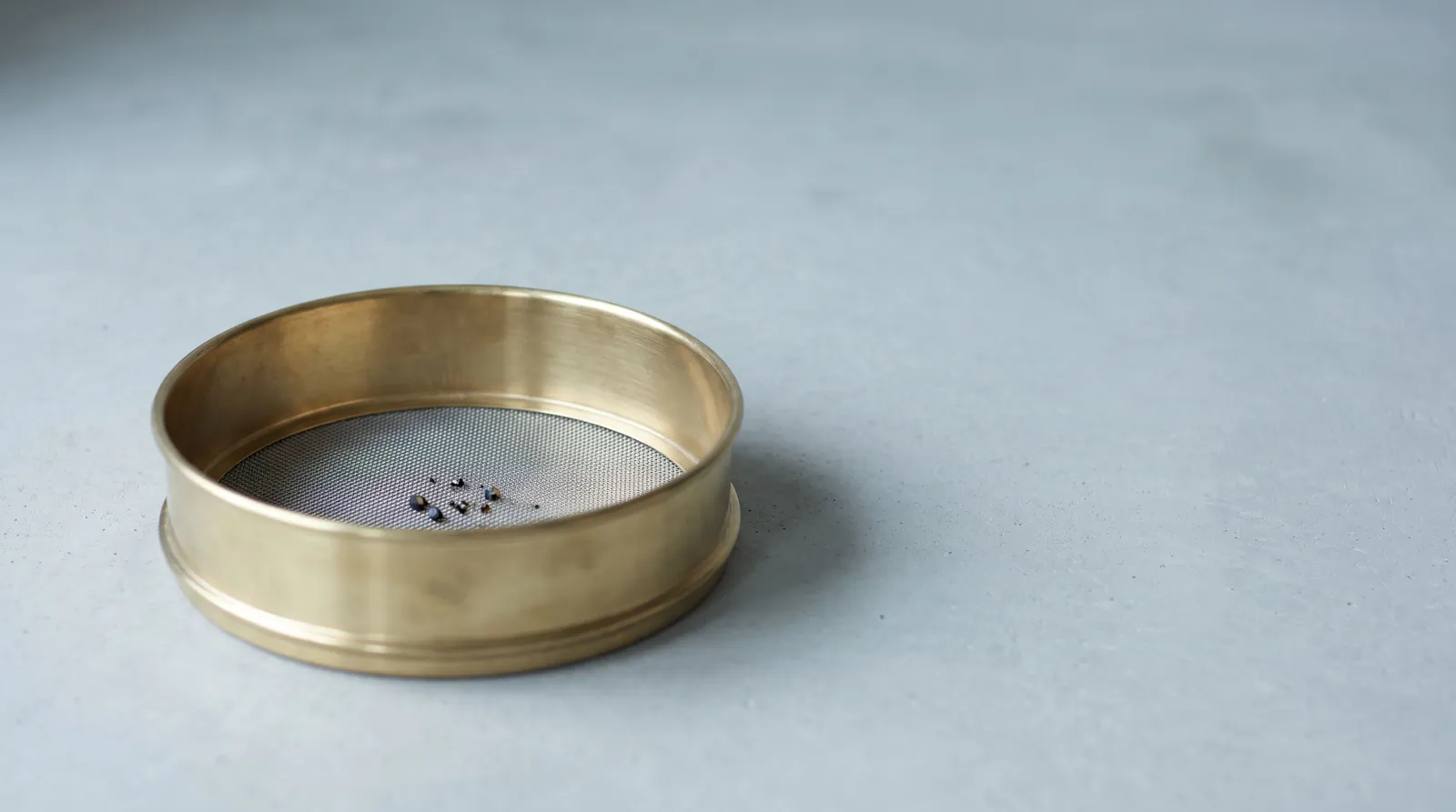 A brass sieve holding a few dark grains on a pale grey surface