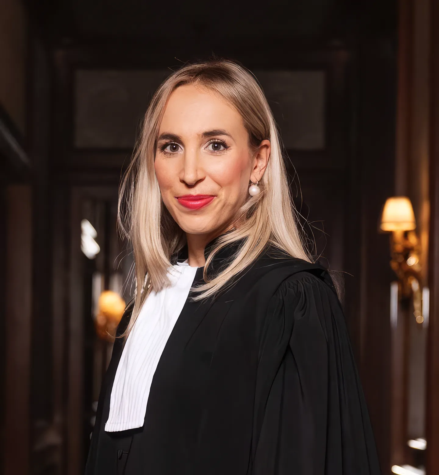 Femme blonde en robe noire d'avocat souriante dans un intérieur aux murs sombres.