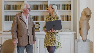 Un homme âgé et une femme souriante discutent dans une bibliothèque avec un ordinateur portable.