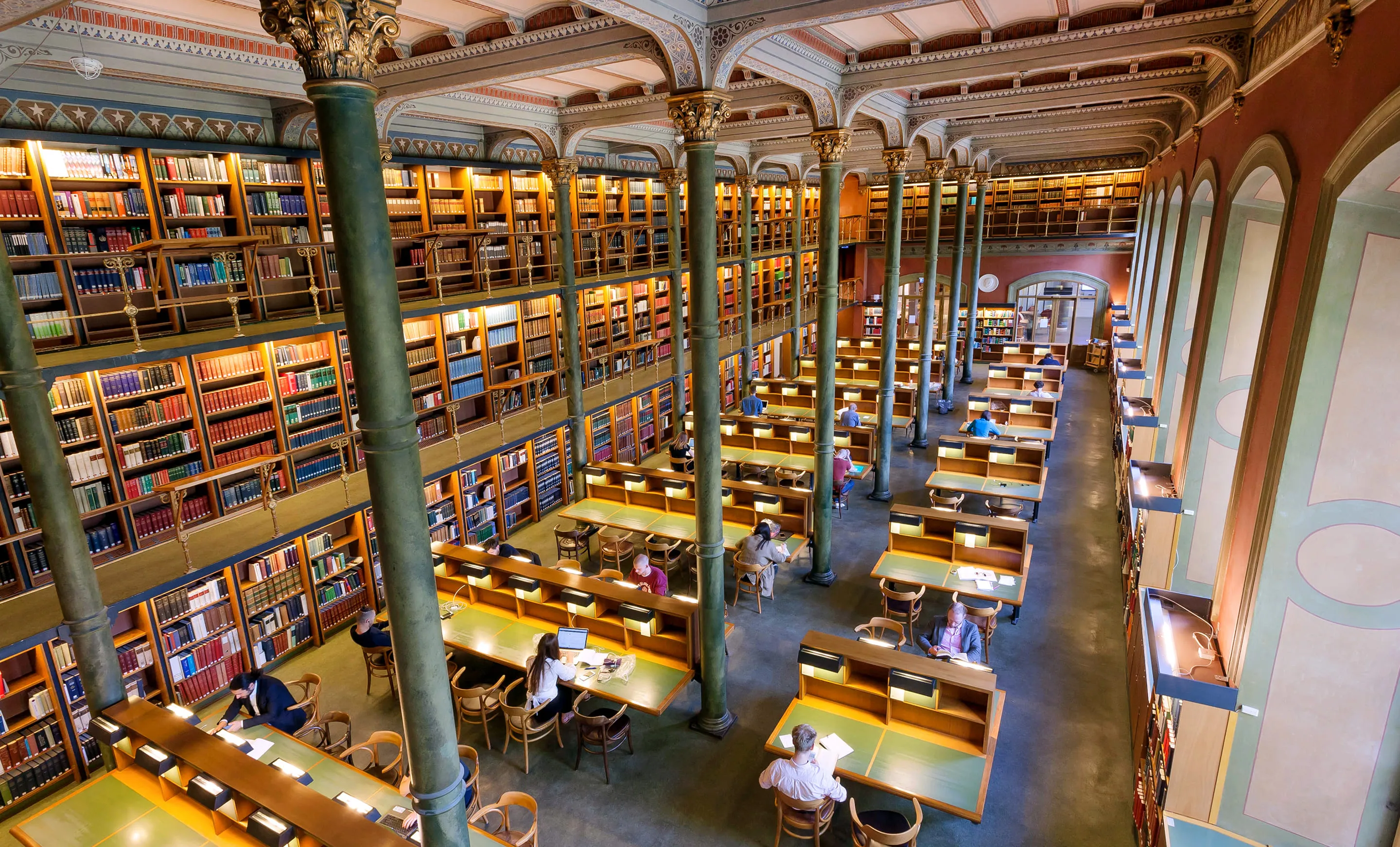 Grande bibliothèque historique avec des étagères en bois remplies de livres, des colonnes décoratives, des tables d'étude individuelles éclairées et des personnes travaillant ou lisant.