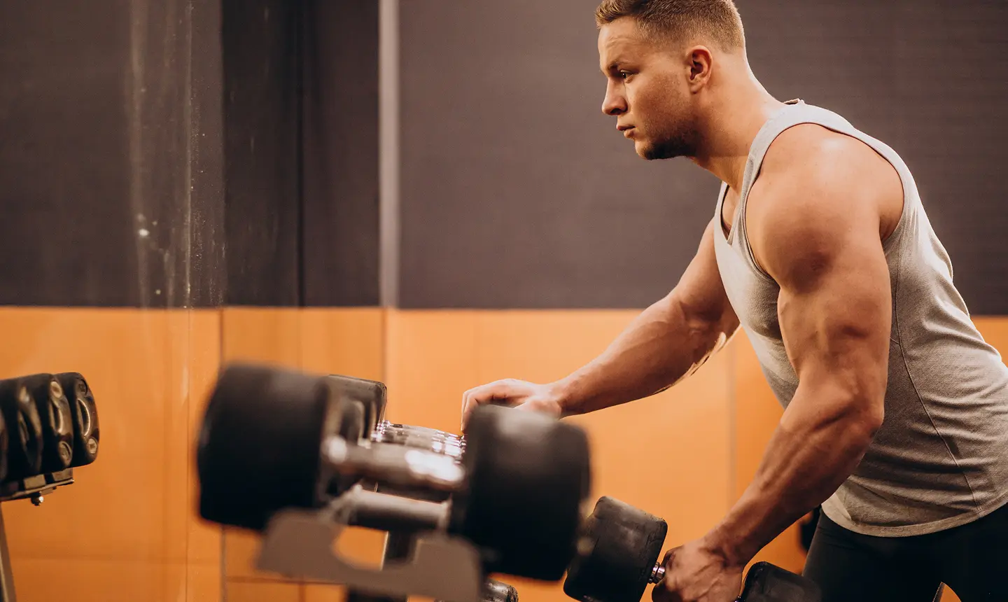 Man in gray tank top lifting dumbbells in a gym.