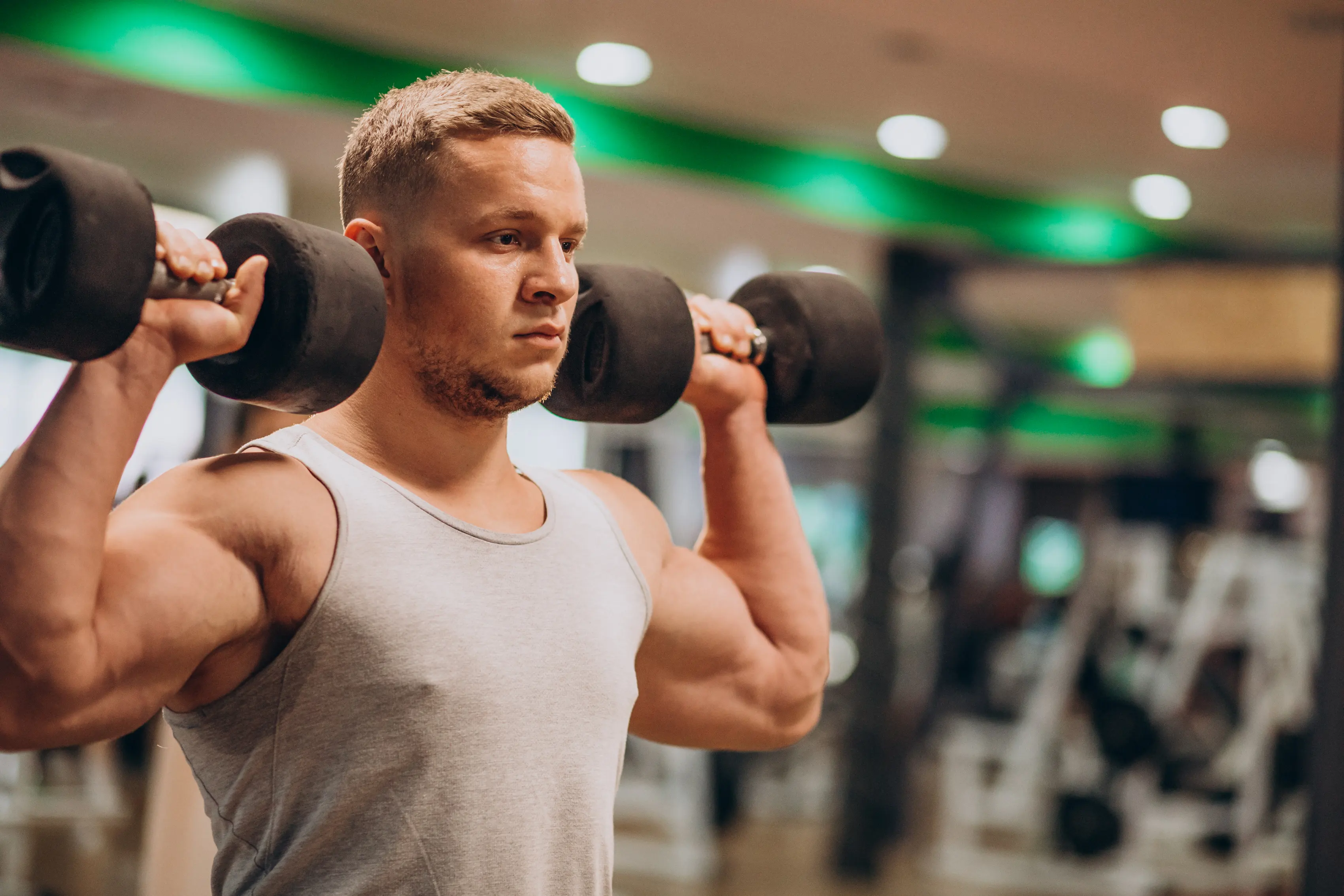Muscular man in a gray tank top lifting black dumbbells in a gym.