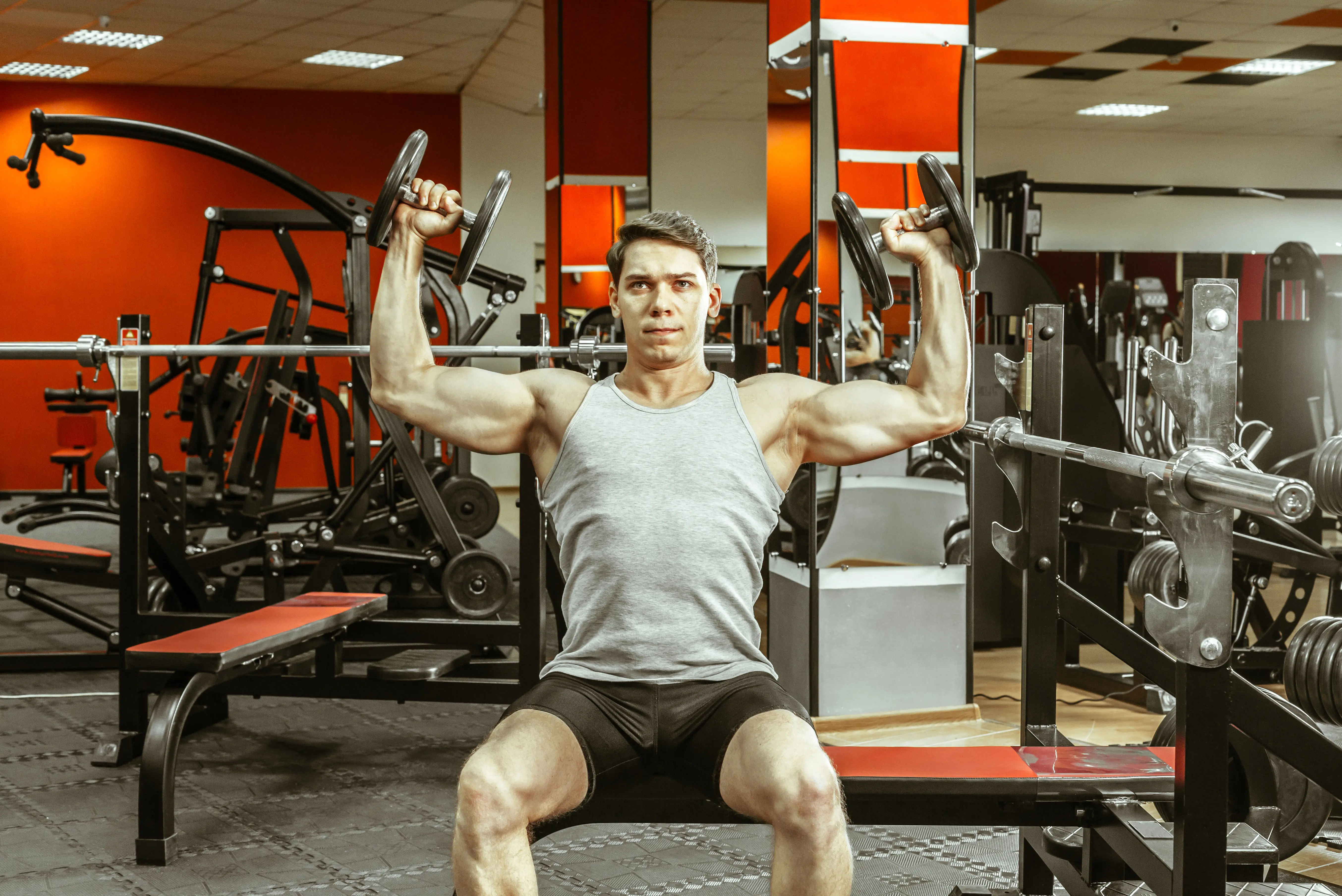 Man in a gray tank top lifting dumbbells while seated on a bench in a gym with red and black workout equipment.