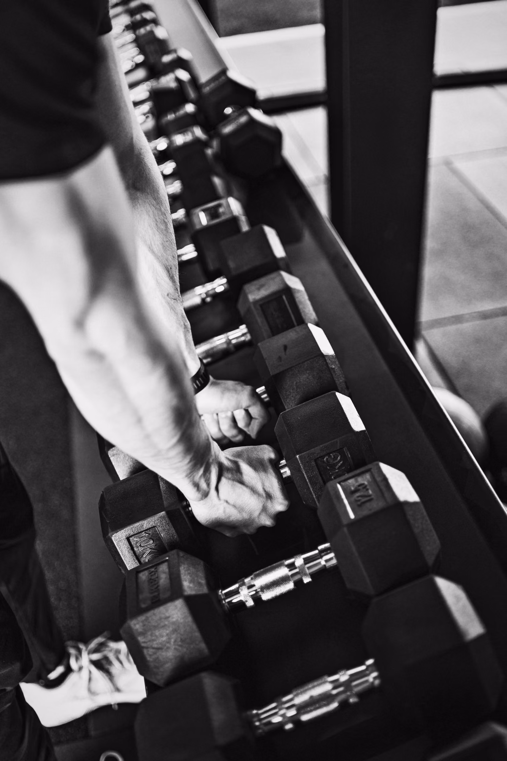 Black and white close-up of a person’s muscular arm and hands gripping a 10kg hexagonal dumbbell from a rack of various weights in a gym.