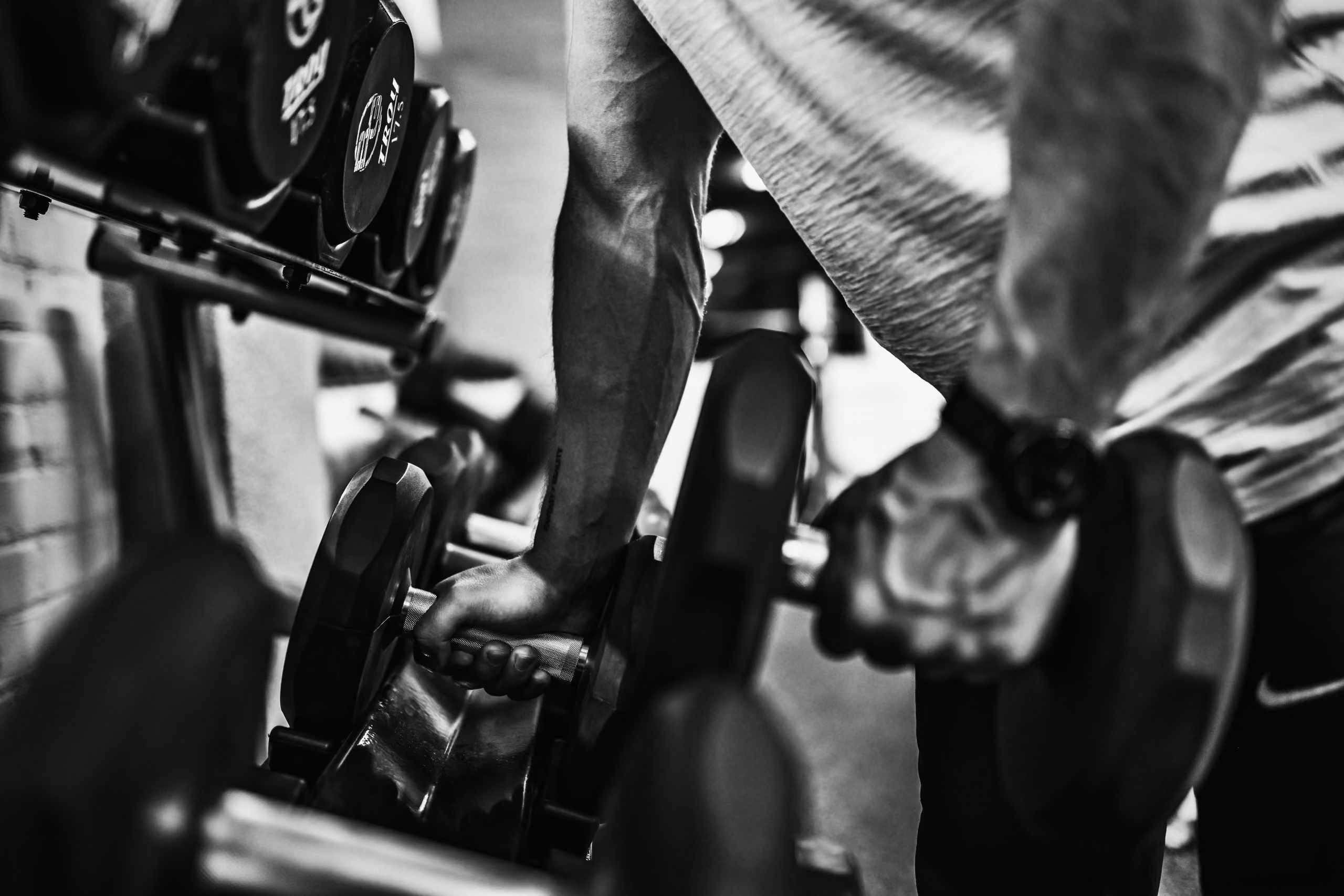Close-up black and white photo of a person grabbing a dumbbell from a rack in a gym, showing muscular forearm and grip.