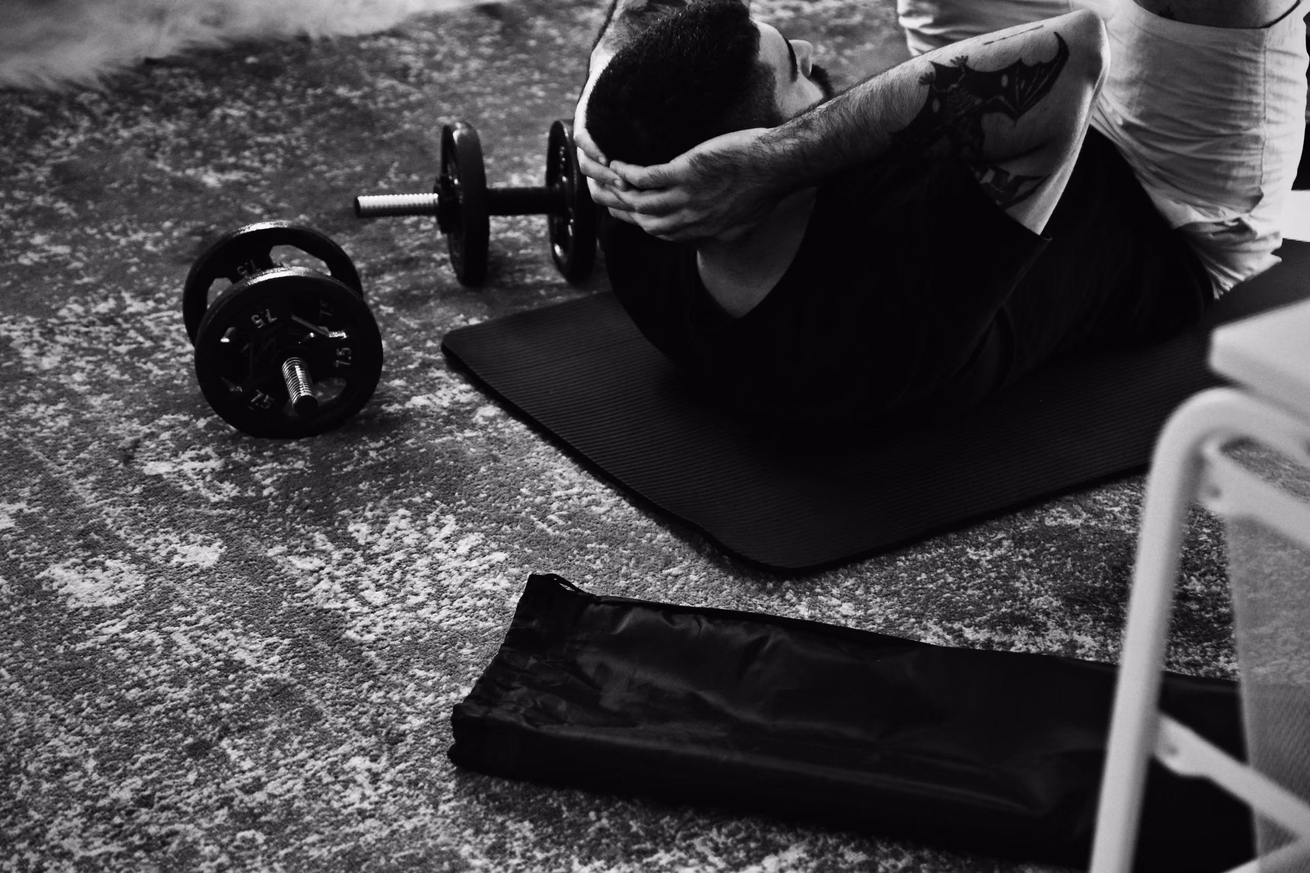 Man with a bat tattoo on his arm doing sit-ups on a black exercise mat beside two dumbbells on a textured floor, seen in black and white.