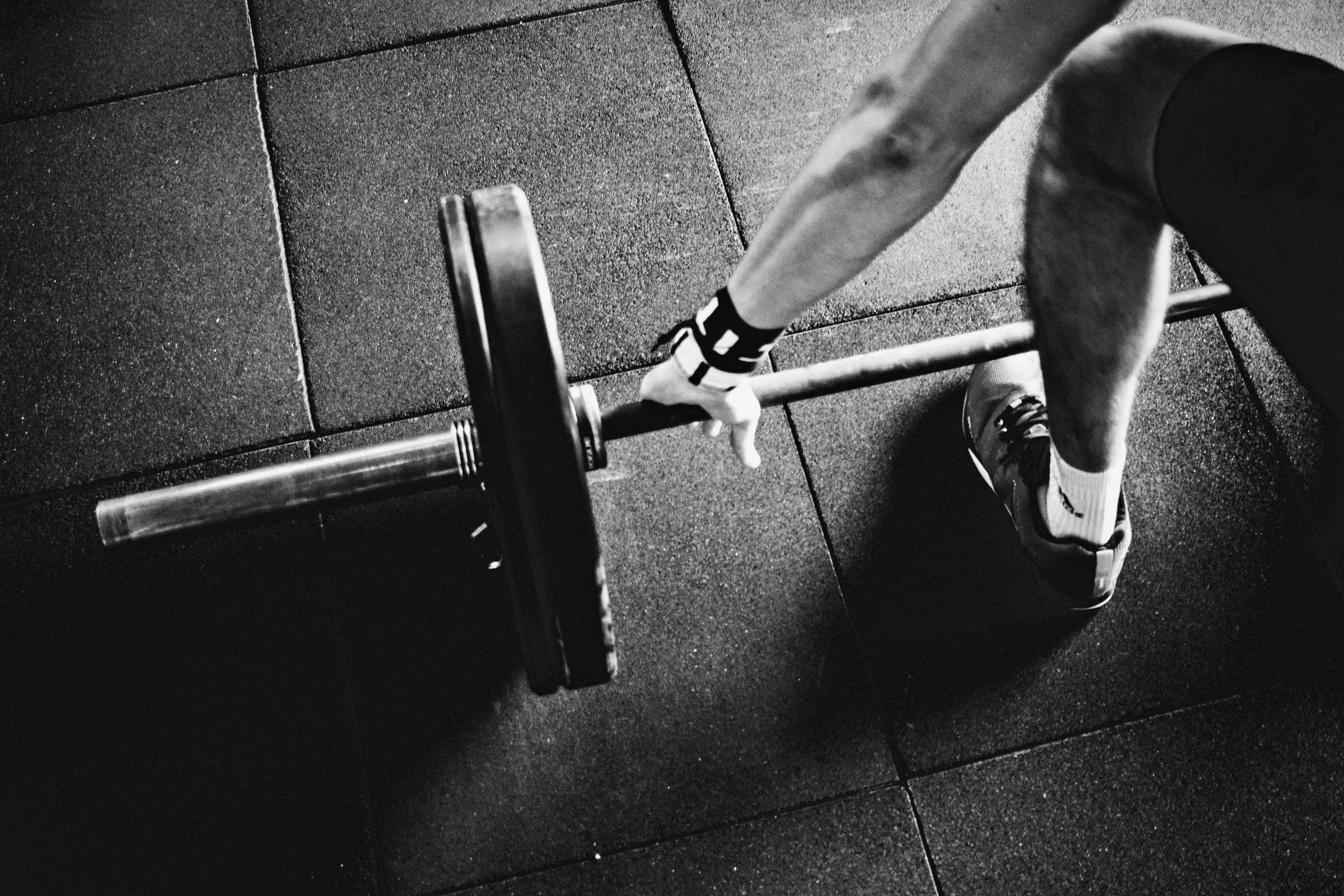 Black and white photo of a person gripping a loaded barbell on a gym floor with rubber mats, wearing a wrist wrap, athletic shoes, and socks.