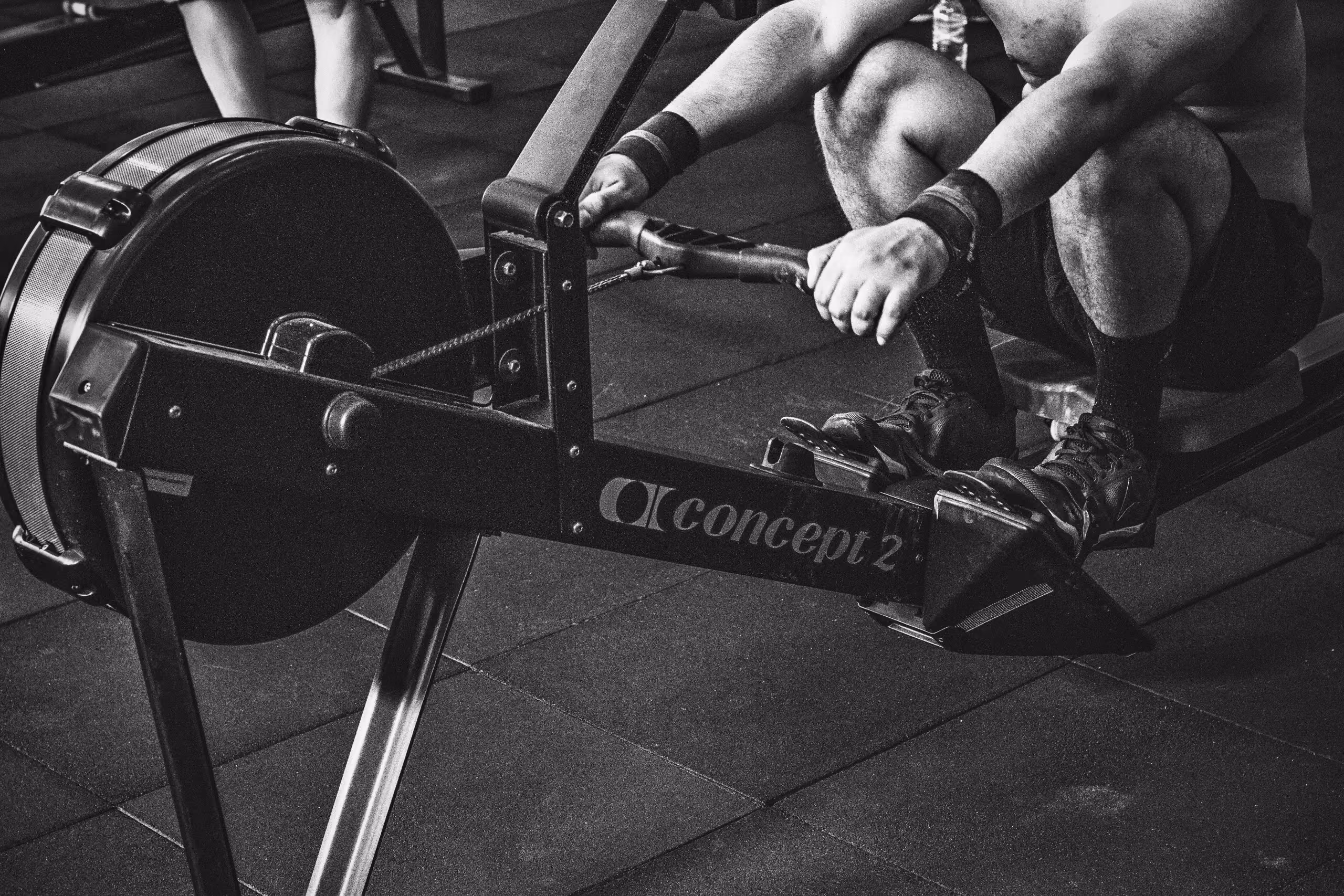Black and white close-up of a person using a Concept2 rowing machine, gripping the handle with feet strapped on the footrests.