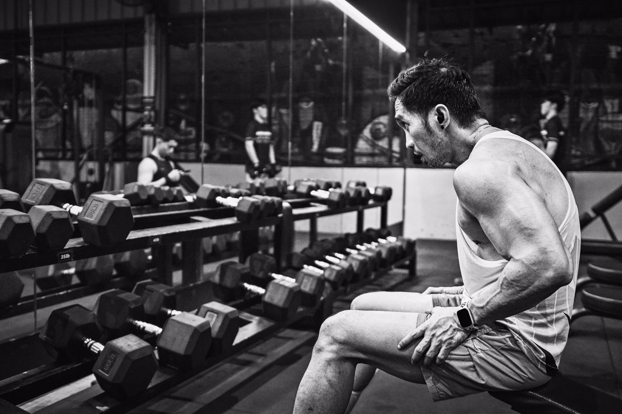 Muscular man in a sleeveless shirt sitting on a gym bench, resting and looking towards dumbbells on racks in front of him in a gym with other people in the background.