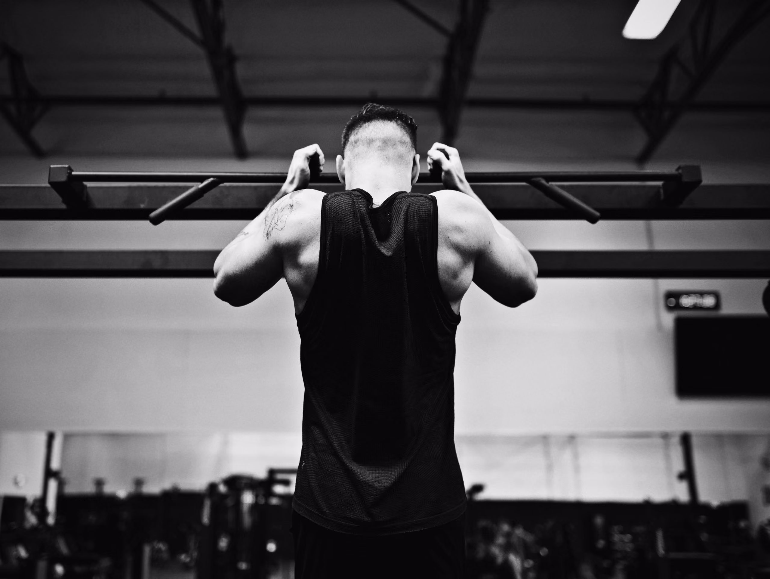A muscular man seen from behind doing pull-ups on a horizontal bar in a gym with workout equipment and mirrors in the background.
