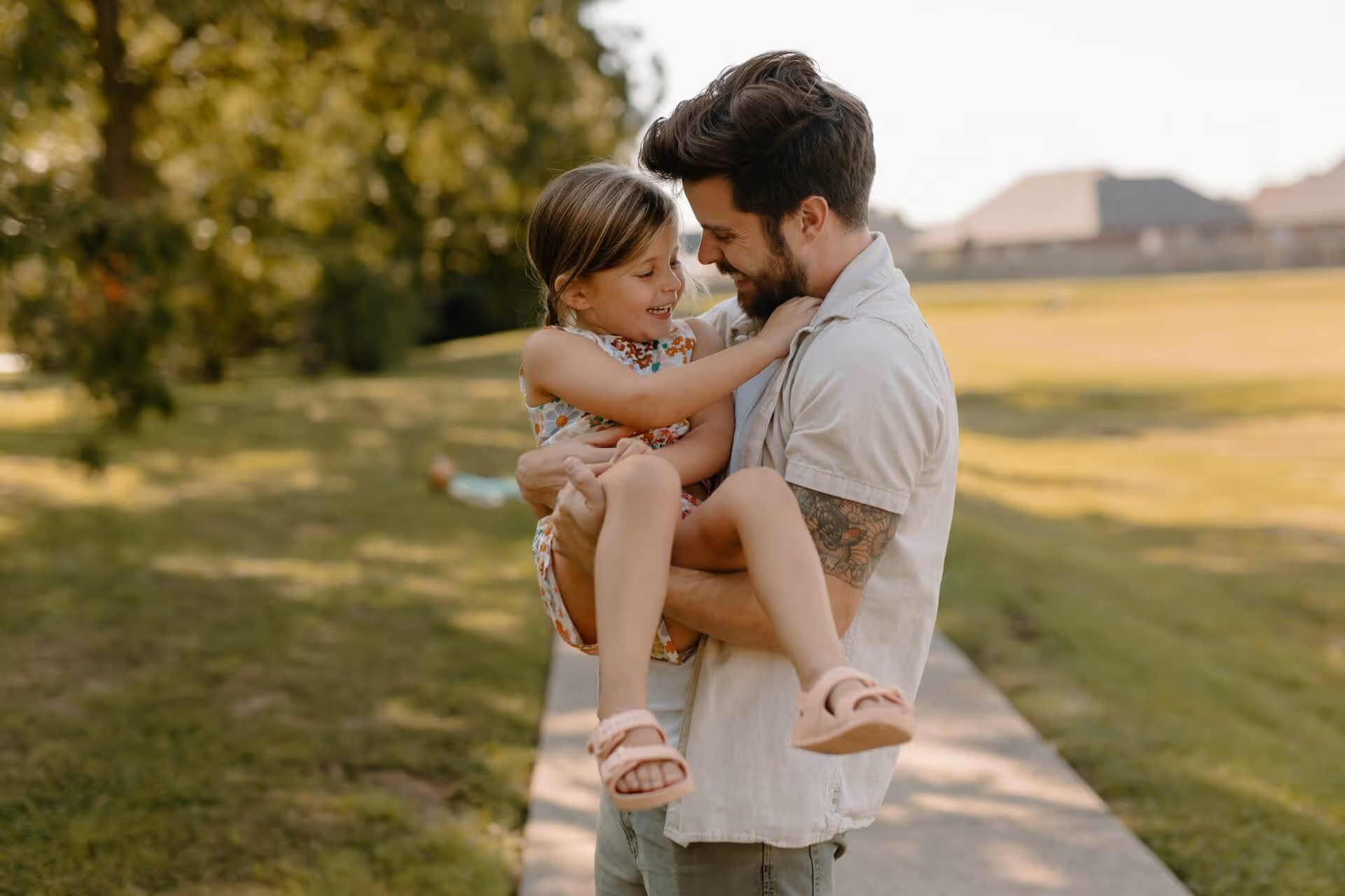 Father smiling and holding his smiling daughter in his arms on a sunlit park path.