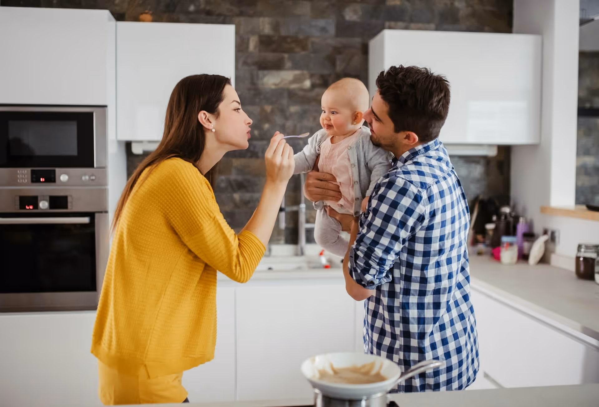 Mother feeding smiling baby held by father in modern kitchen with stone wall and white cabinets.