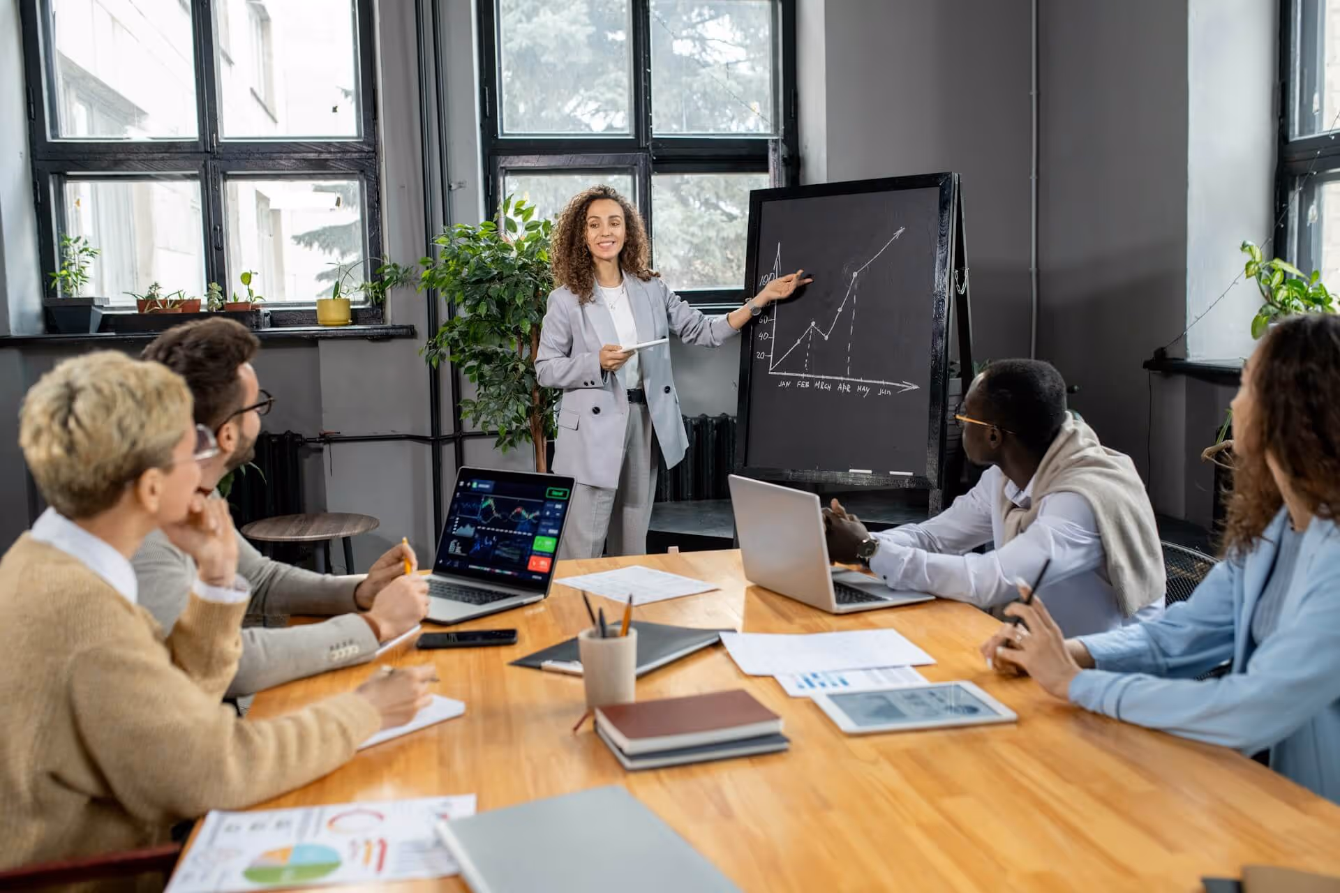 Businesswoman pointing to a rising graph on a blackboard in a meeting with four colleagues.