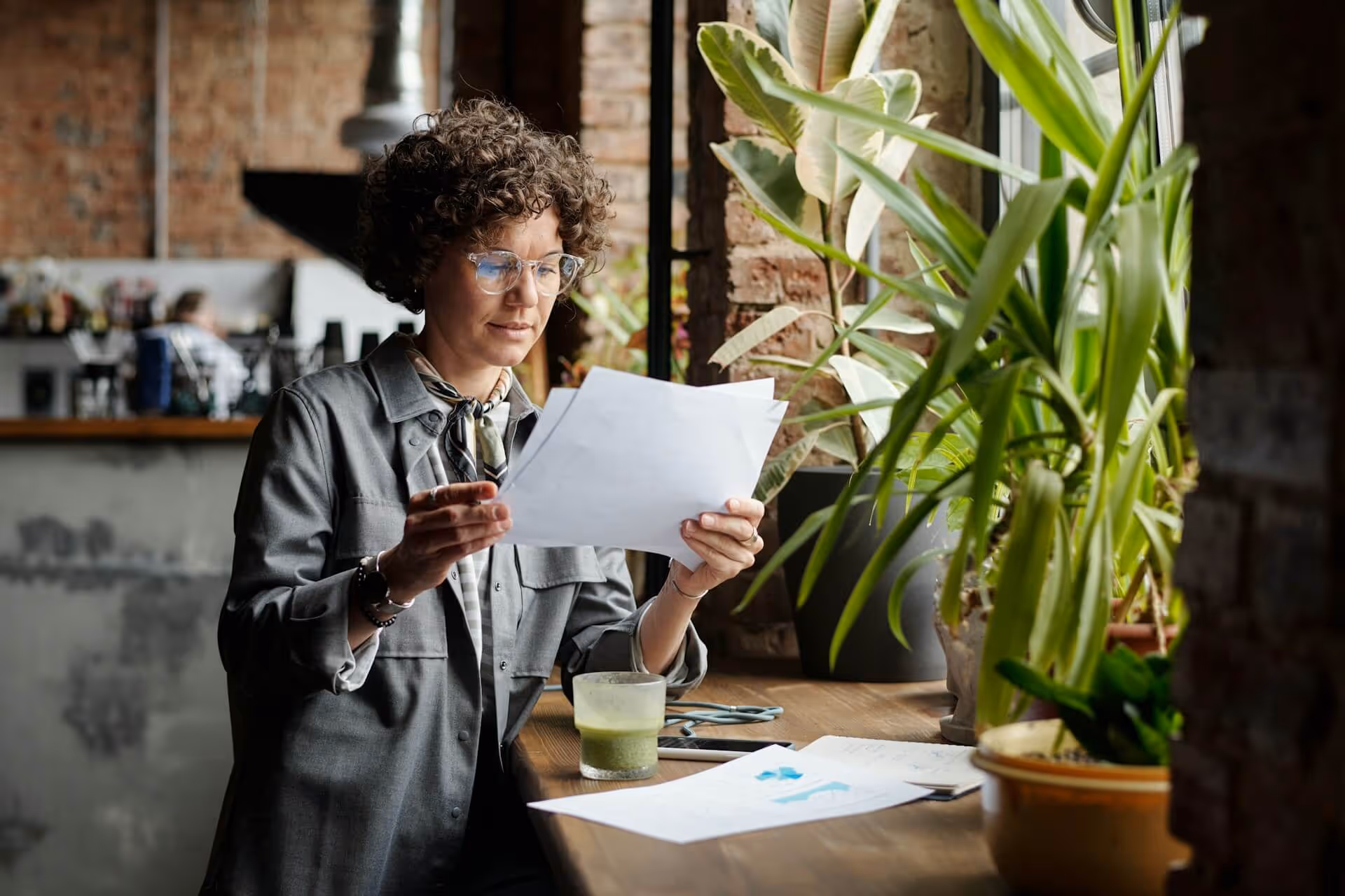 Person reading papers at a wooden counter by a window with green plants and a green smoothie nearby.