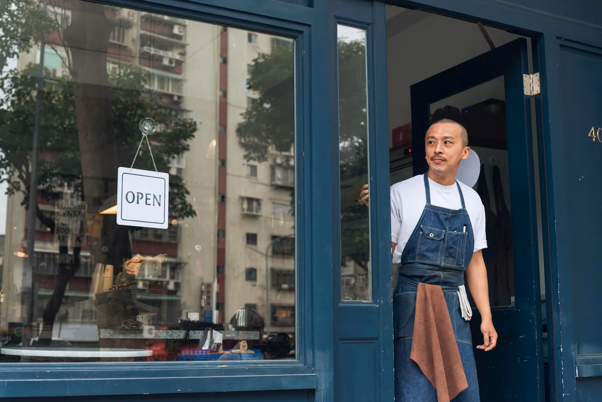 Man in a denim apron standing at the open door of a blue storefront with an open sign.
