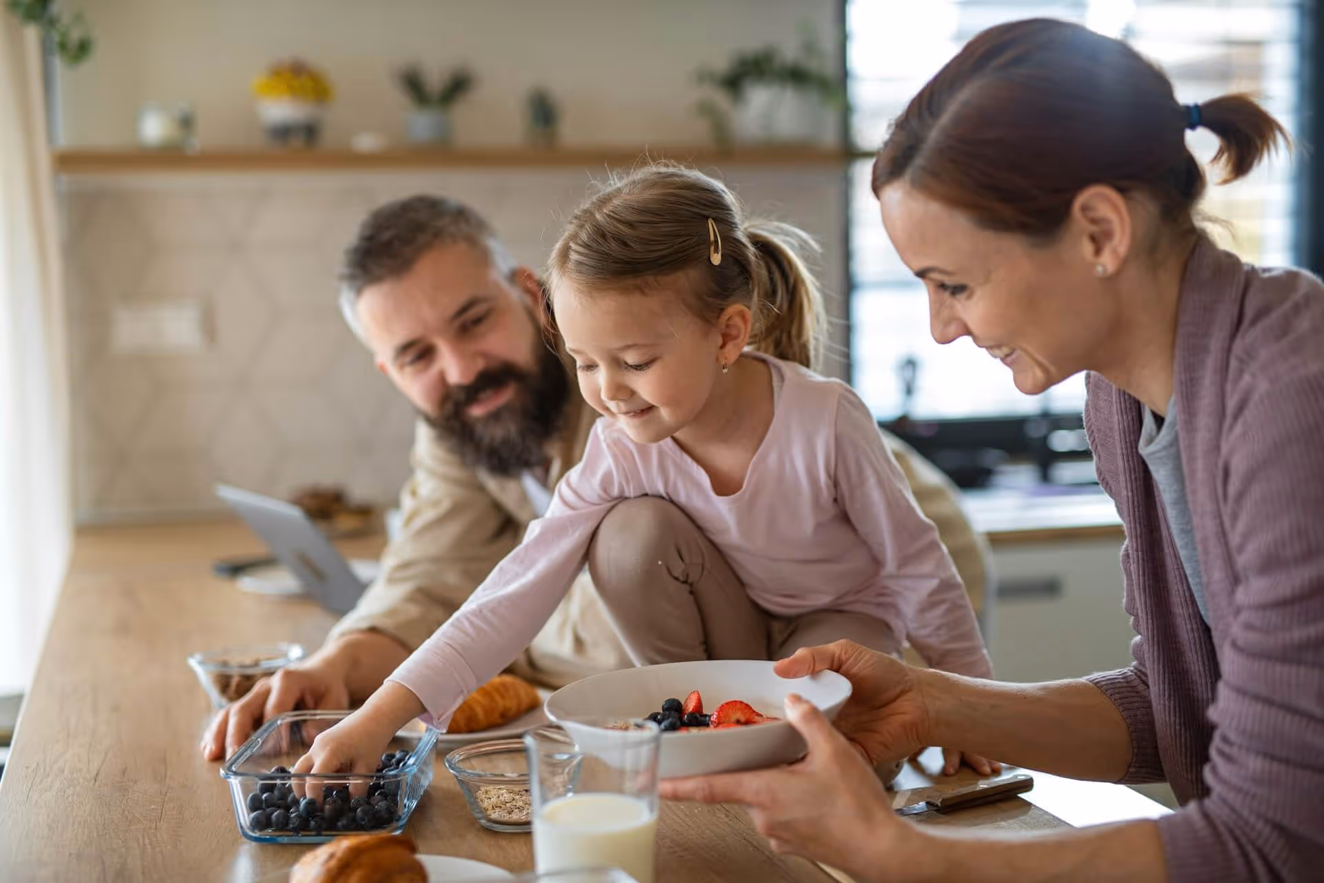 Smiling parents helping daughter pick blueberries for breakfast bowl in a cozy kitchen.