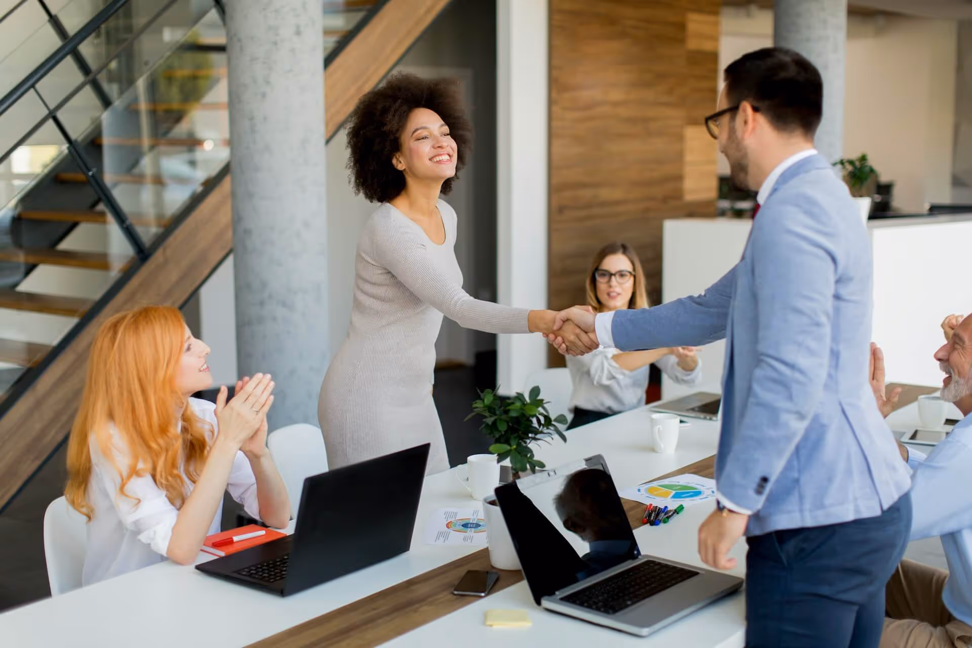 Two colleagues shaking hands across a conference table while others watch and applaud in an office.