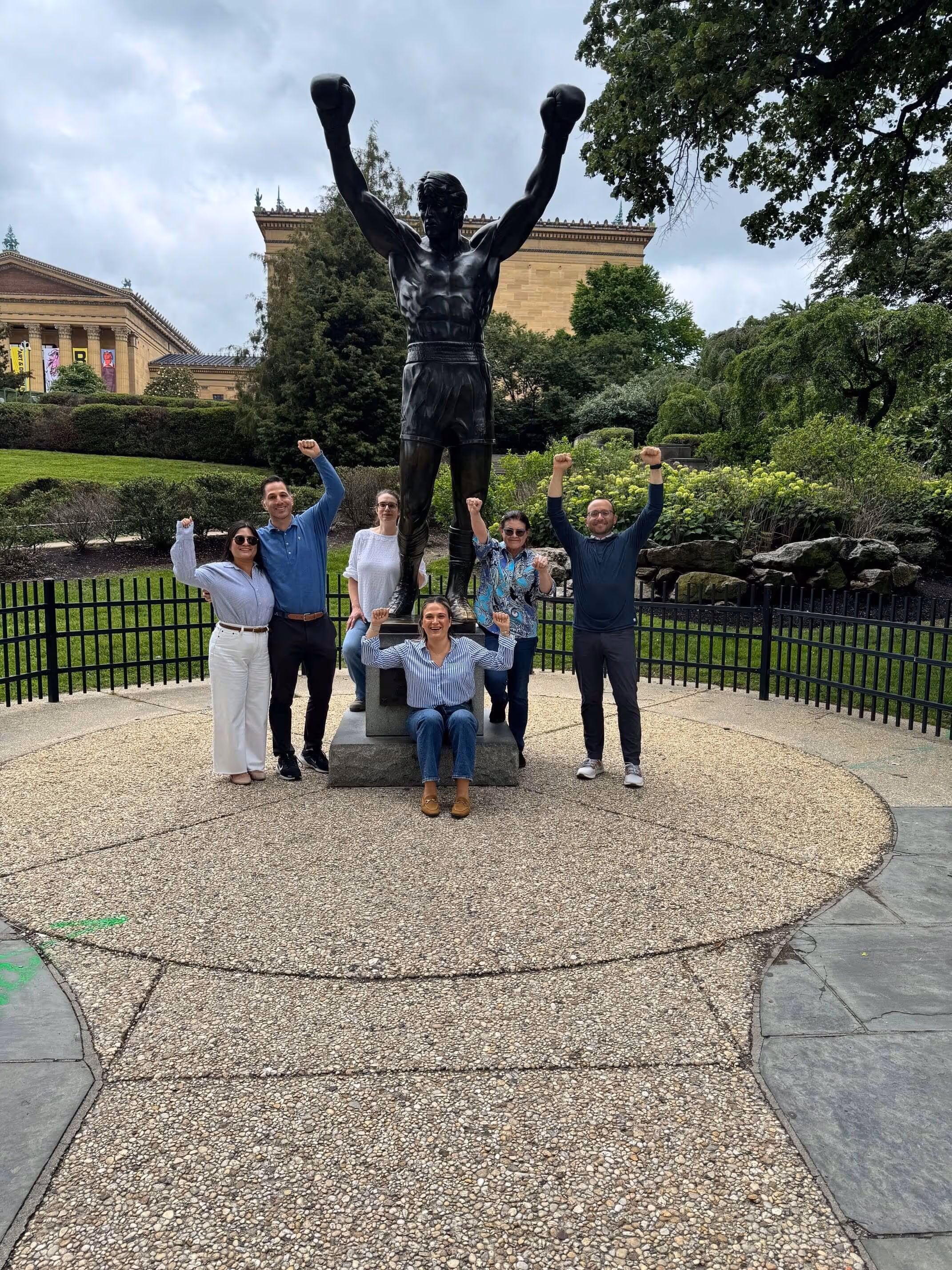 Group of six people posing with raised fists around the Rocky Balboa statue outdoors.