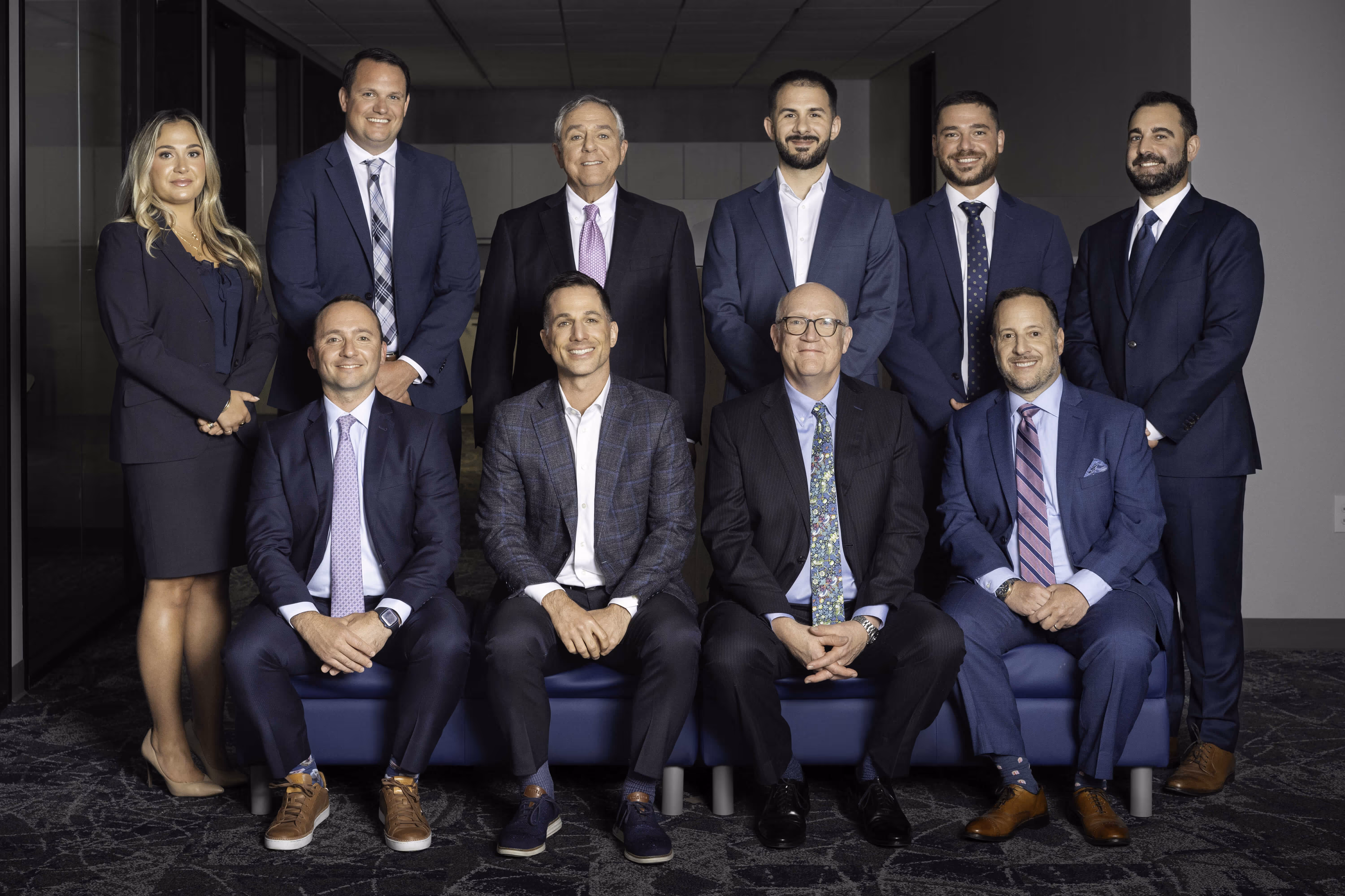 Group photo of 11 professionally dressed men and one woman posing indoors on and behind a bench.