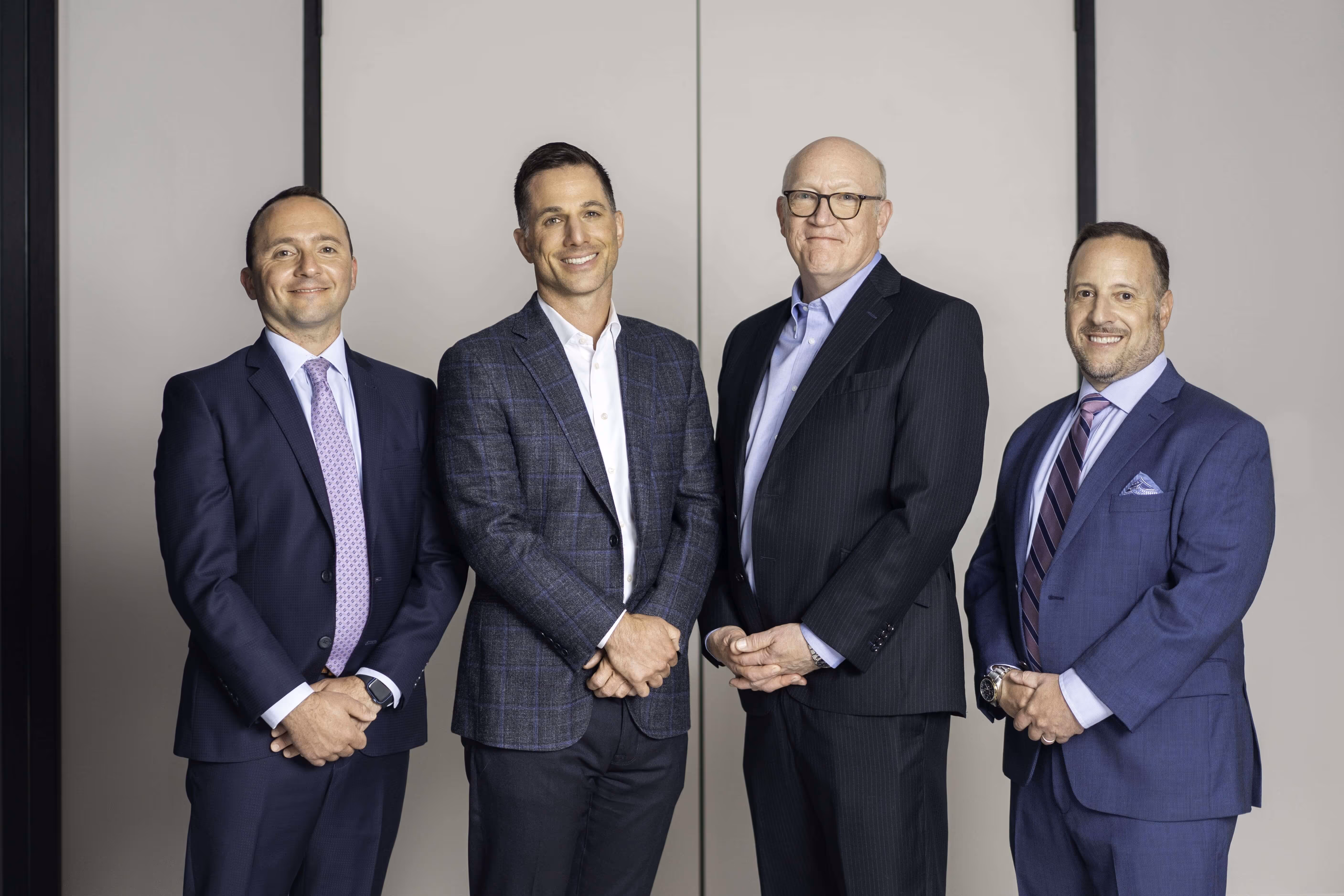 Four businessmen in suits standing and smiling against a plain light background.