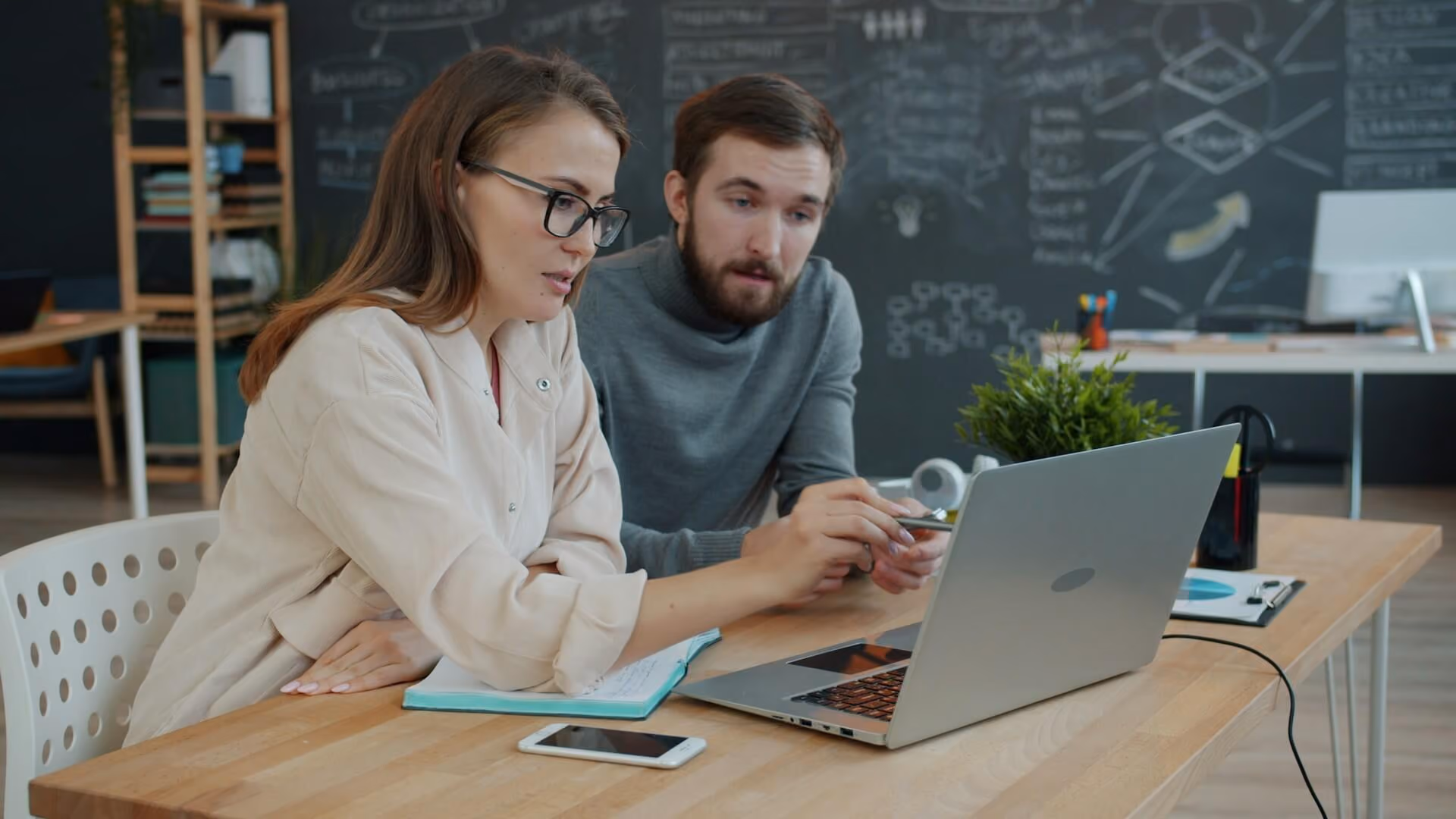 Two colleagues sit at a desk, focused on a laptop screen in a modern office space.