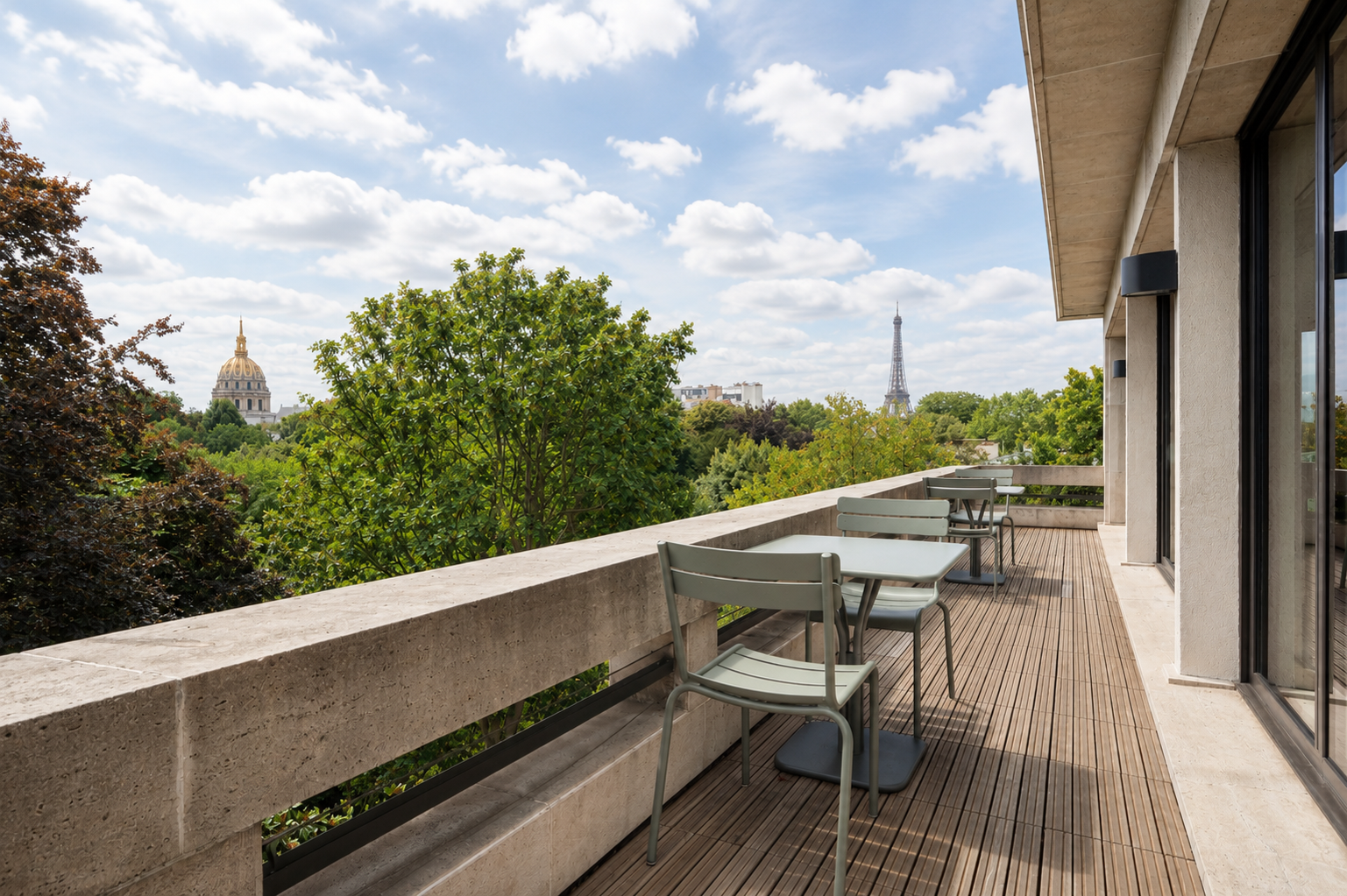 Outdoor terrace with light green tables and chairs on wooden flooring, overlooking lush green trees with a clear sky and the Eiffel Tower and a dome-topped building visible in the distance.