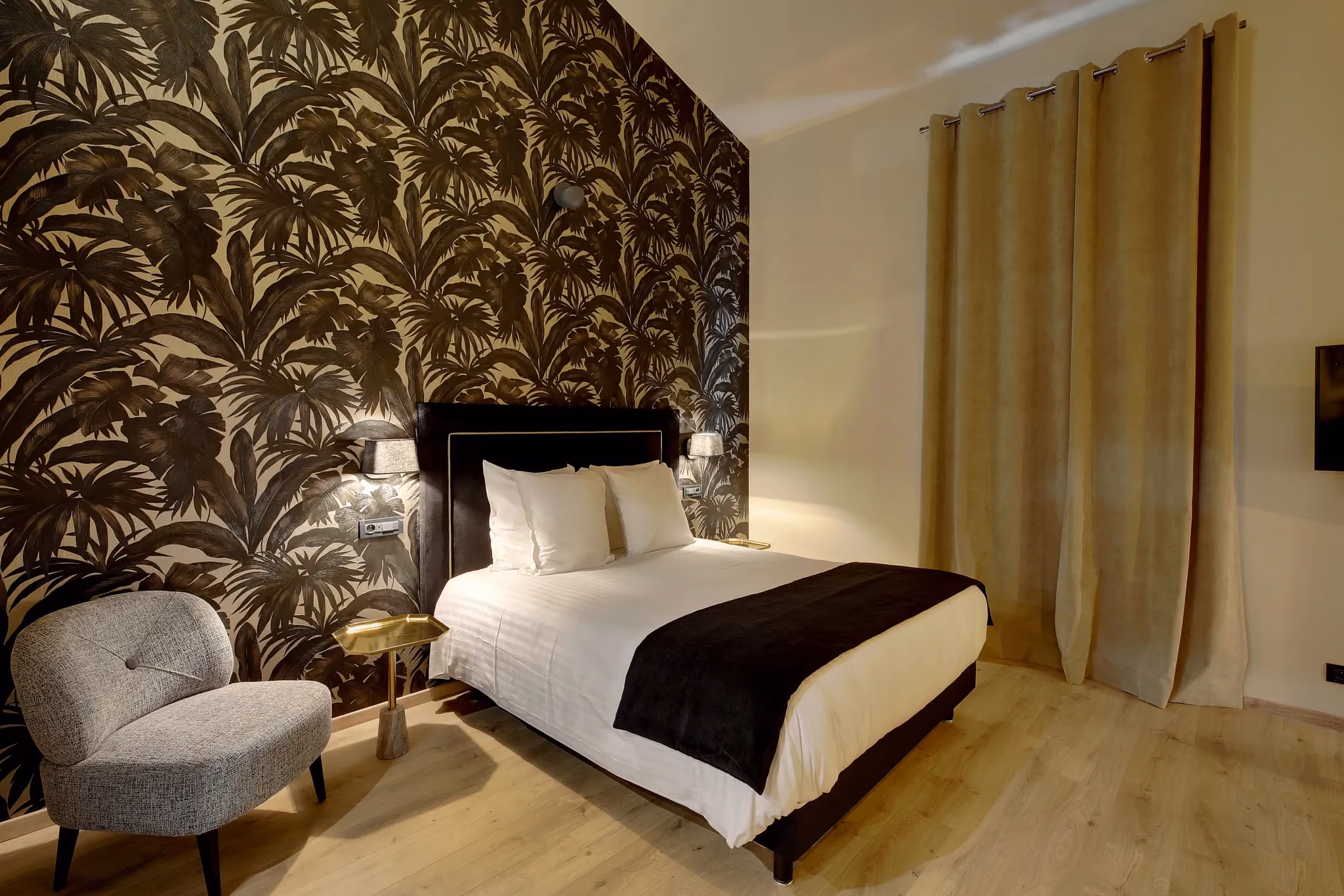 Chambre avec grand lit blanc, couverture noire, fauteuil gris et mur avec papier peint feuilles.