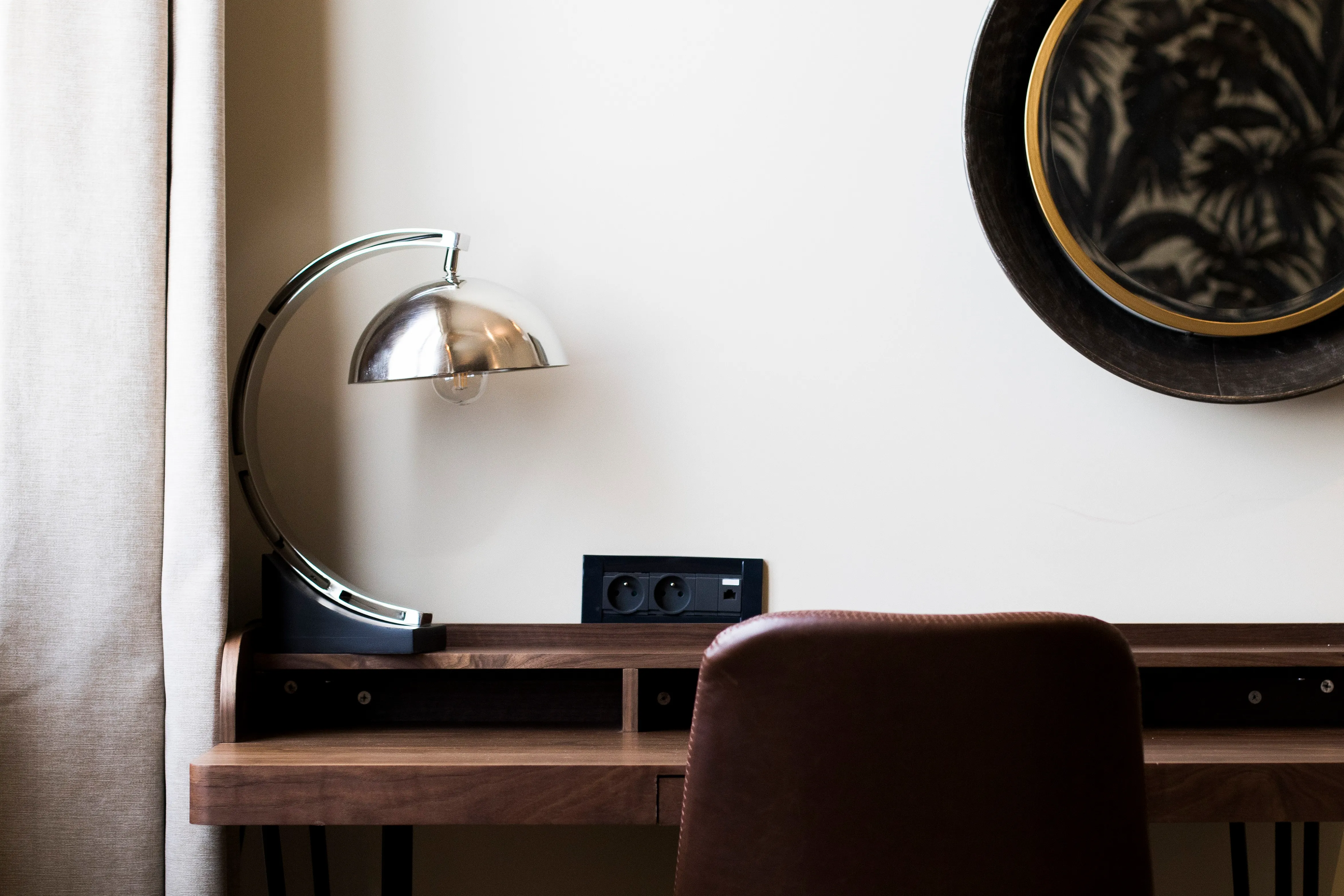 Lampe de bureau chromée sur un bureau en bois avec une chaise marron et un miroir rond.