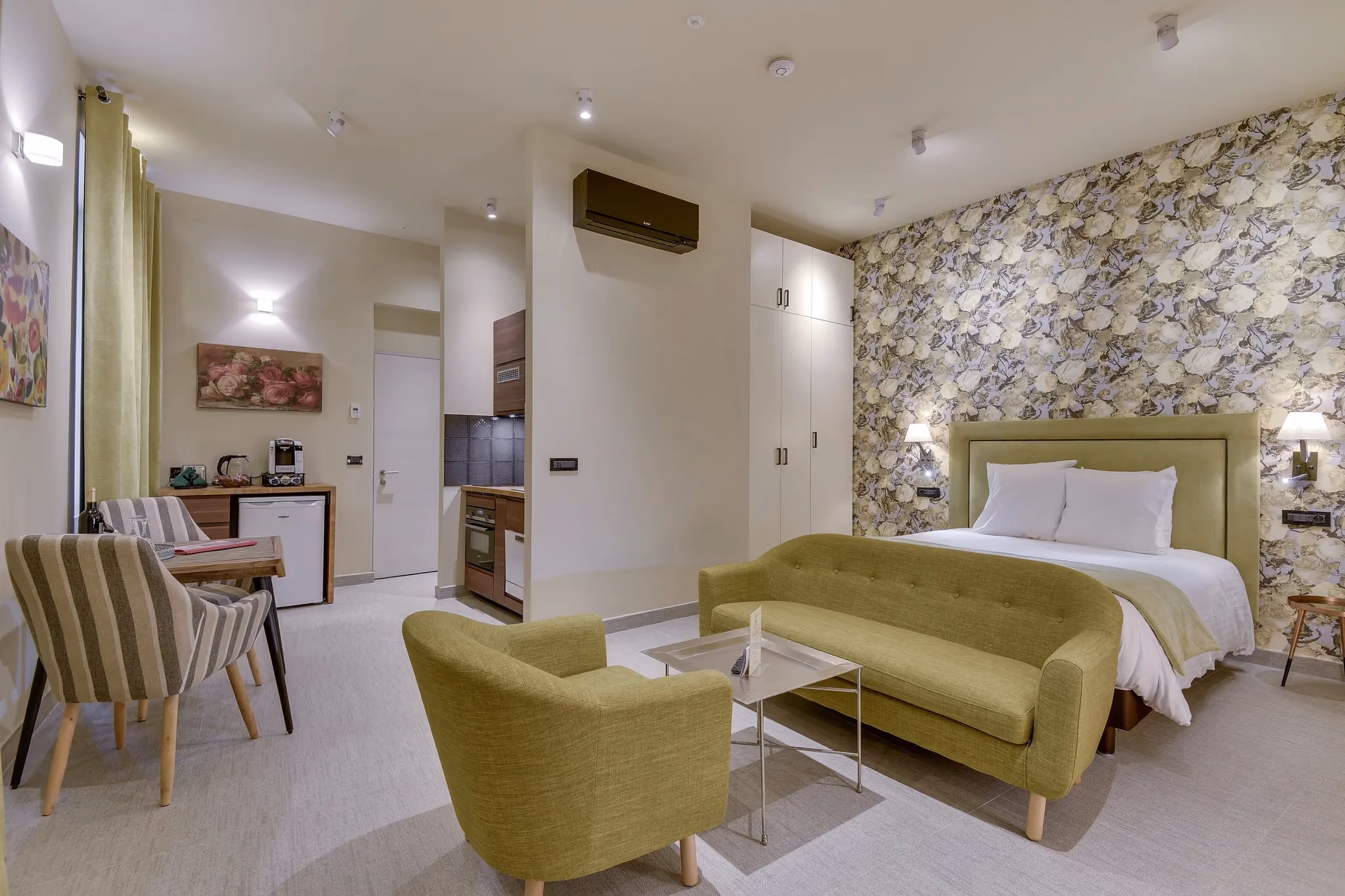 Chambre moderne avec lit double, canapé, fauteuils verts, table basse, coin cuisine et décor floral.