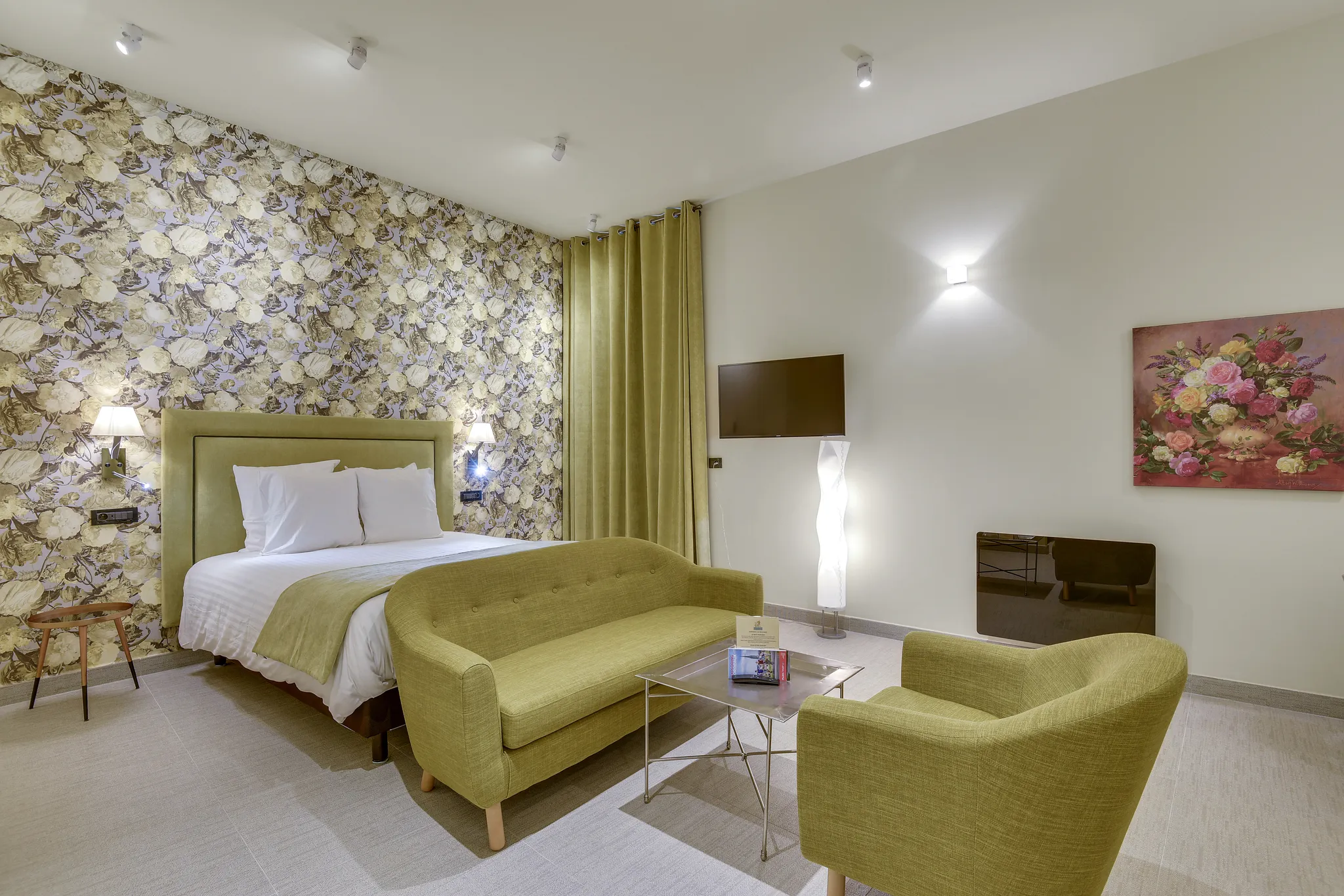 Chambre avec lit double, canapé vert, fauteuil, table basse et mur à papier peint floral.