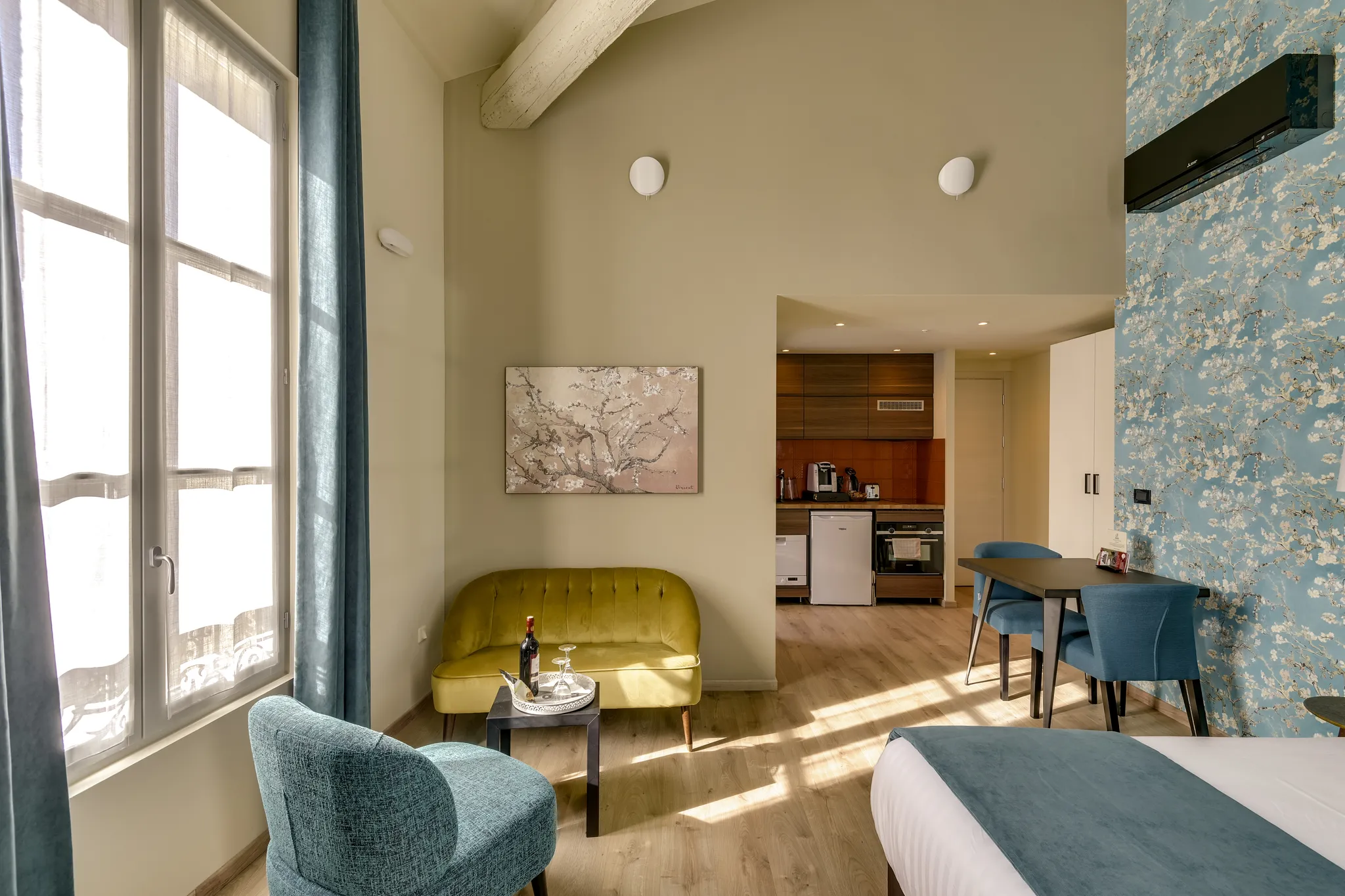 Coin salon lumineux avec fauteuil bleu, canapé vert, table noire et cuisine ouverte au fond.