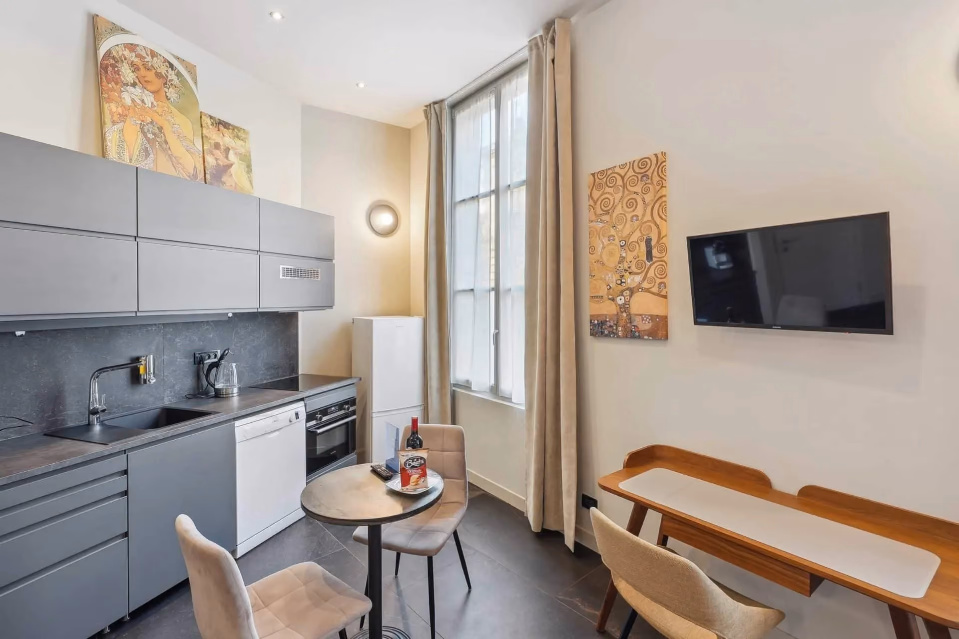 Cuisine moderne grise avec table ronde, chaises, réfrigérateur, four, et télévision murale.