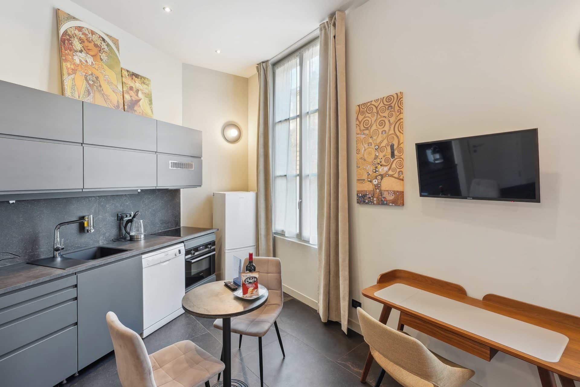 Cuisine moderne grise avec table ronde, chaises, réfrigérateur, four, et télévision murale.