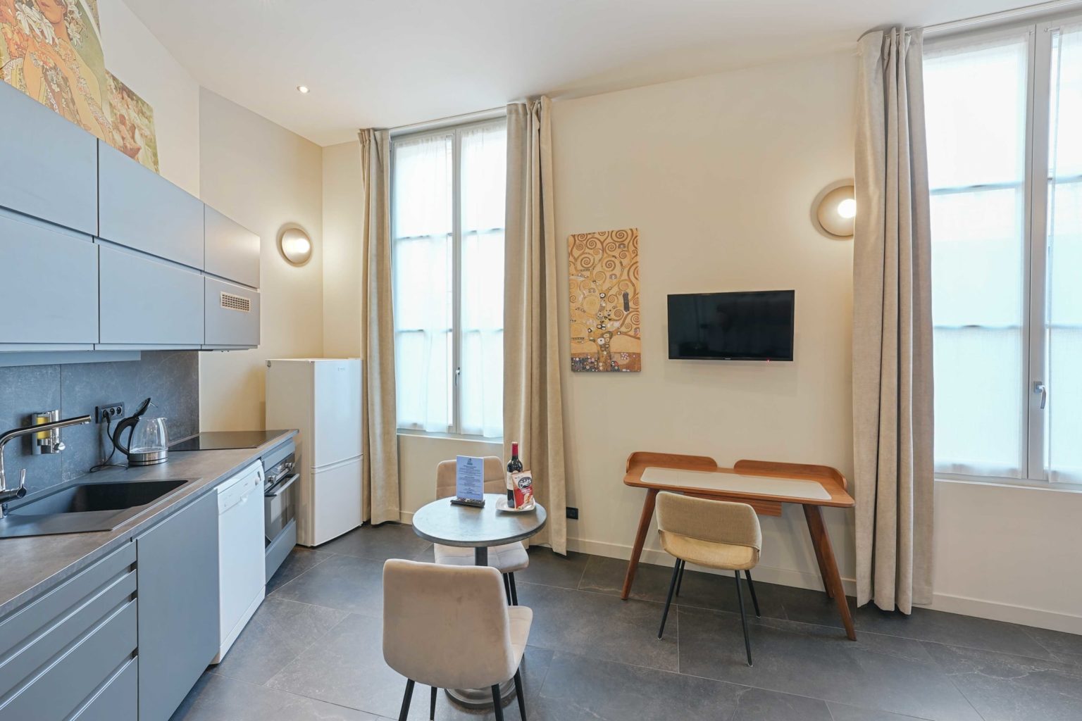 Cuisine moderne avec table ronde, chaises, réfrigérateur blanc et télévision murale dans un appartement clair.